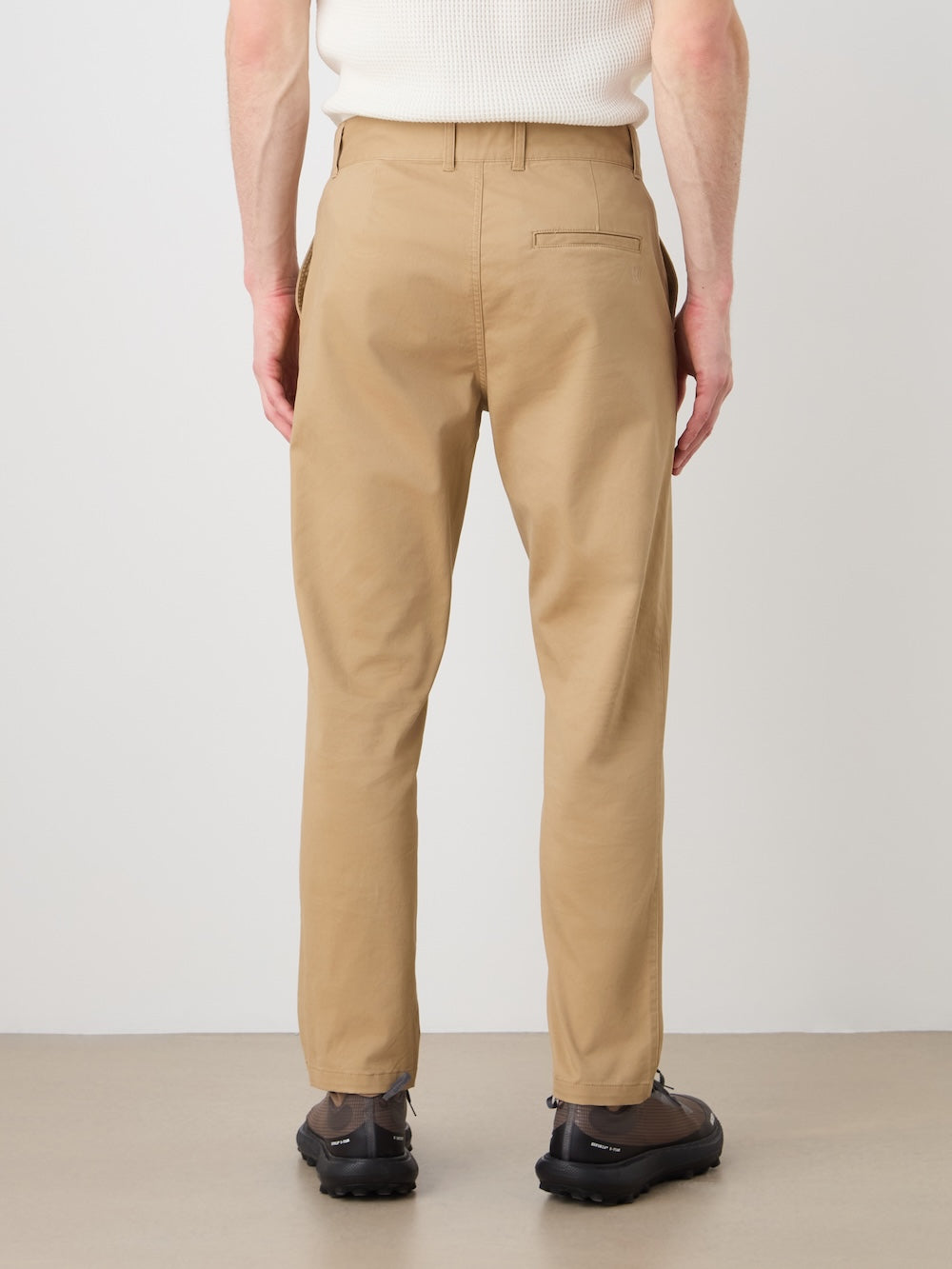 A person wearing We Norwegians Sol Pant Men in tan, a white sleeveless top, and black shoes stands with their back to the camera against a light background.