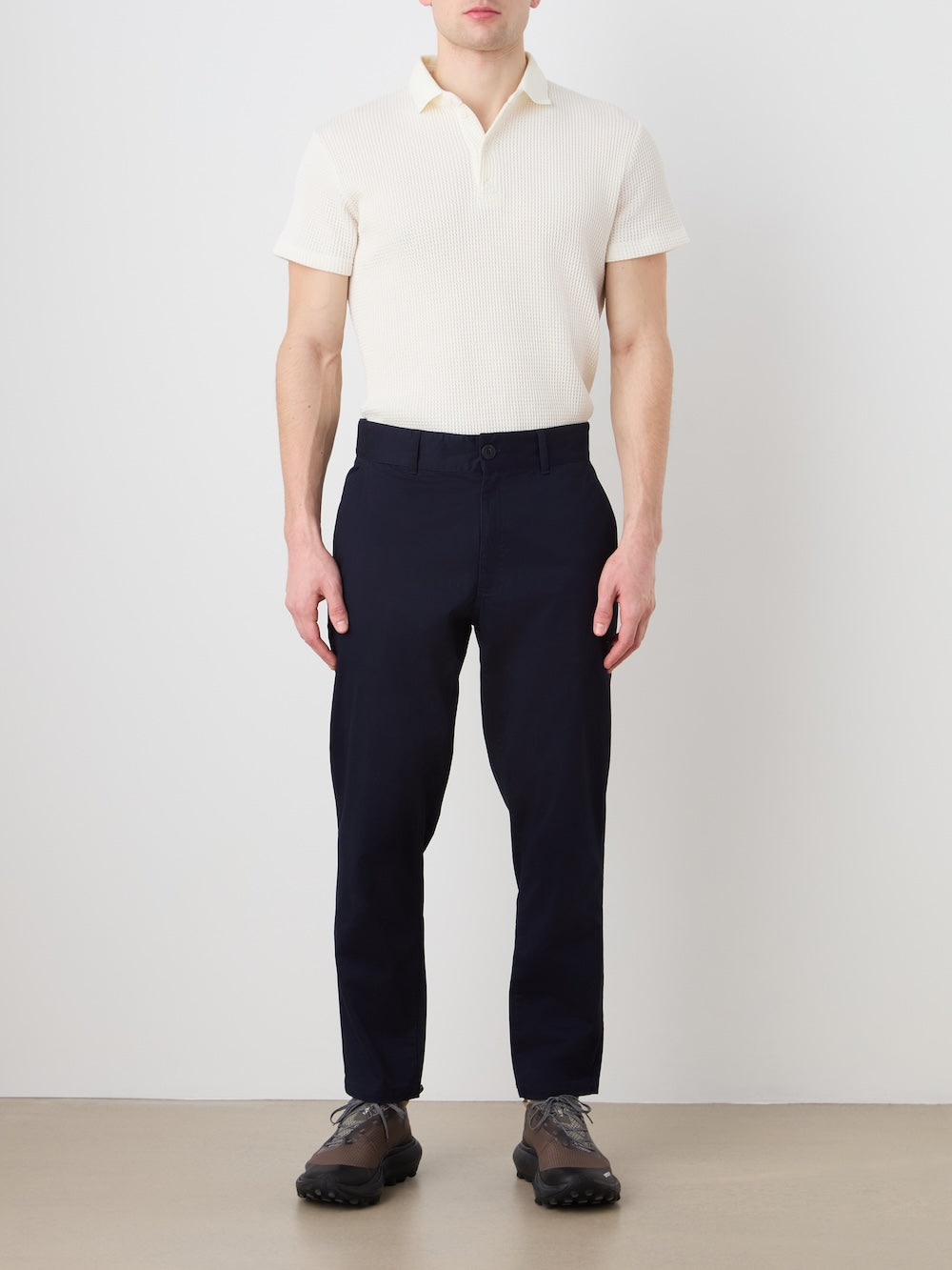 A person stands against a plain background wearing a cream knit polo, We Norwegians Sol Pant Men in dark navy, and brown lace-up shoes. Their arms are relaxed at their sides.