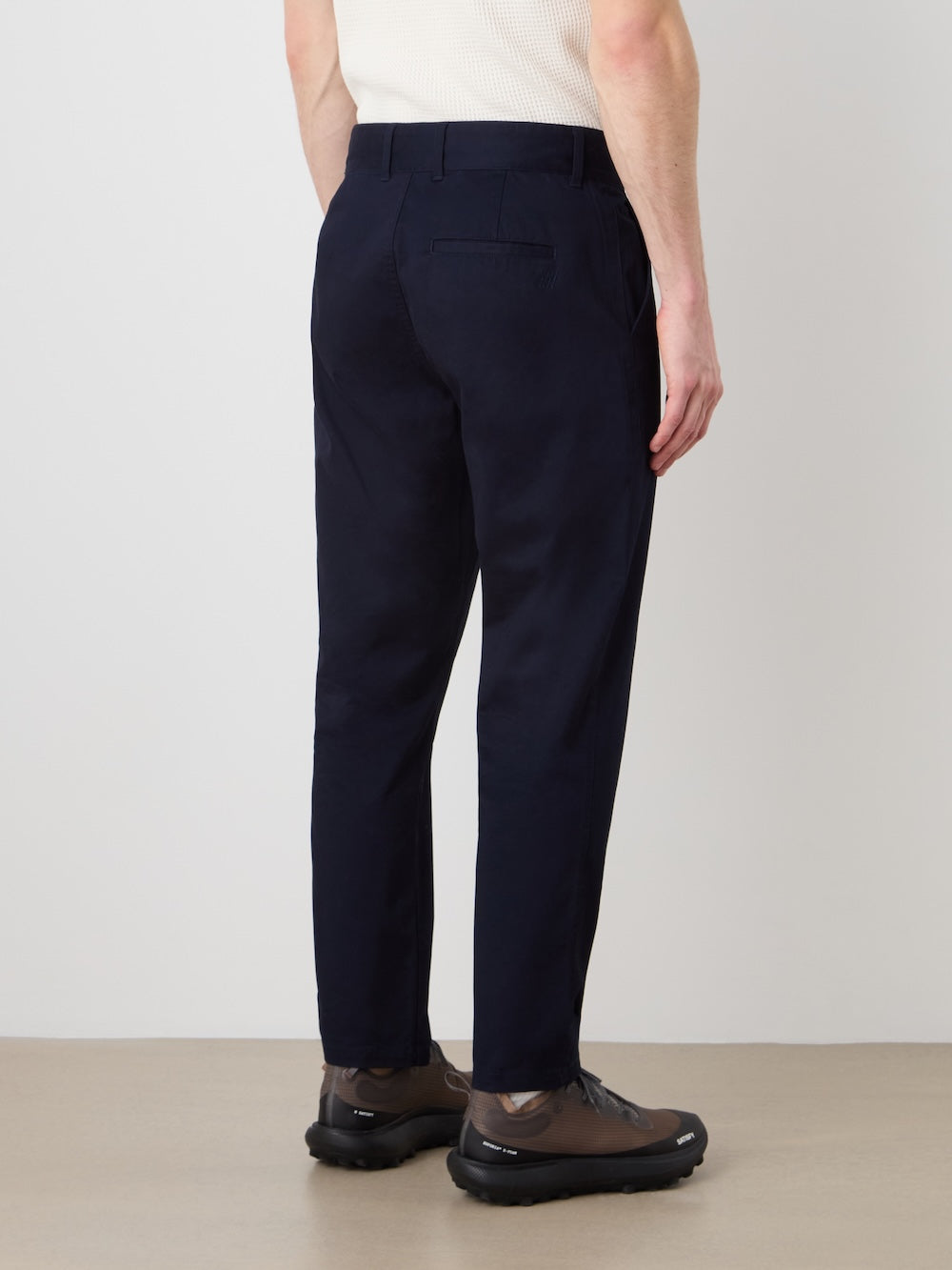 A person stands indoors facing away, wearing We Norwegians Sol Pant Men in dark navy cotton stretch with a tailored fit, paired with brown shoes and a white ribbed shirt against a plain, neutral background.