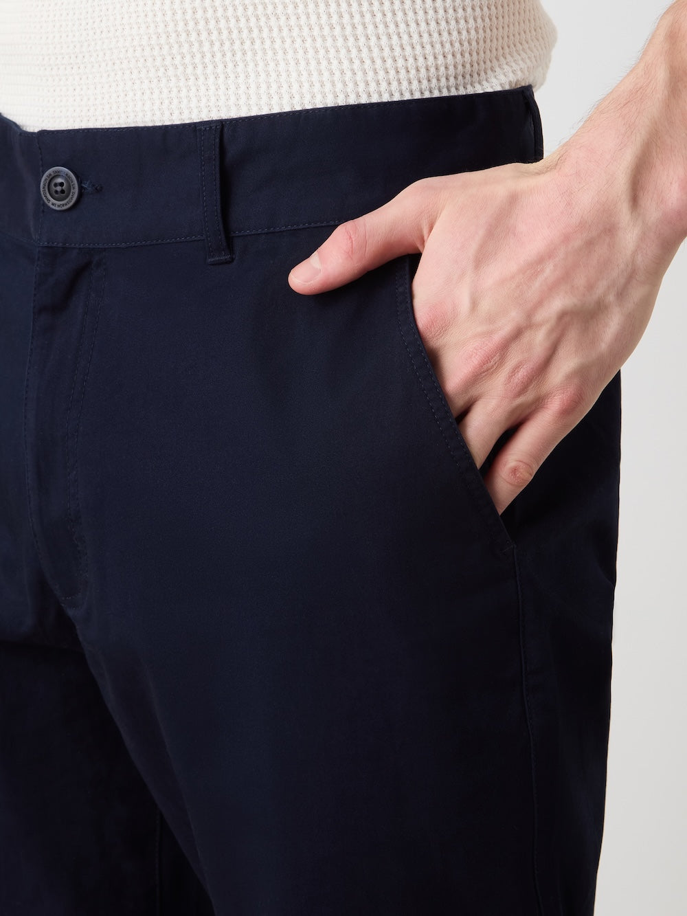 A close-up of a person wearing We Norwegians Sol Pant Men in dark navy cotton stretch, paired with a white textured shirt and their right hand partially tucked into the pant’s front pocket.