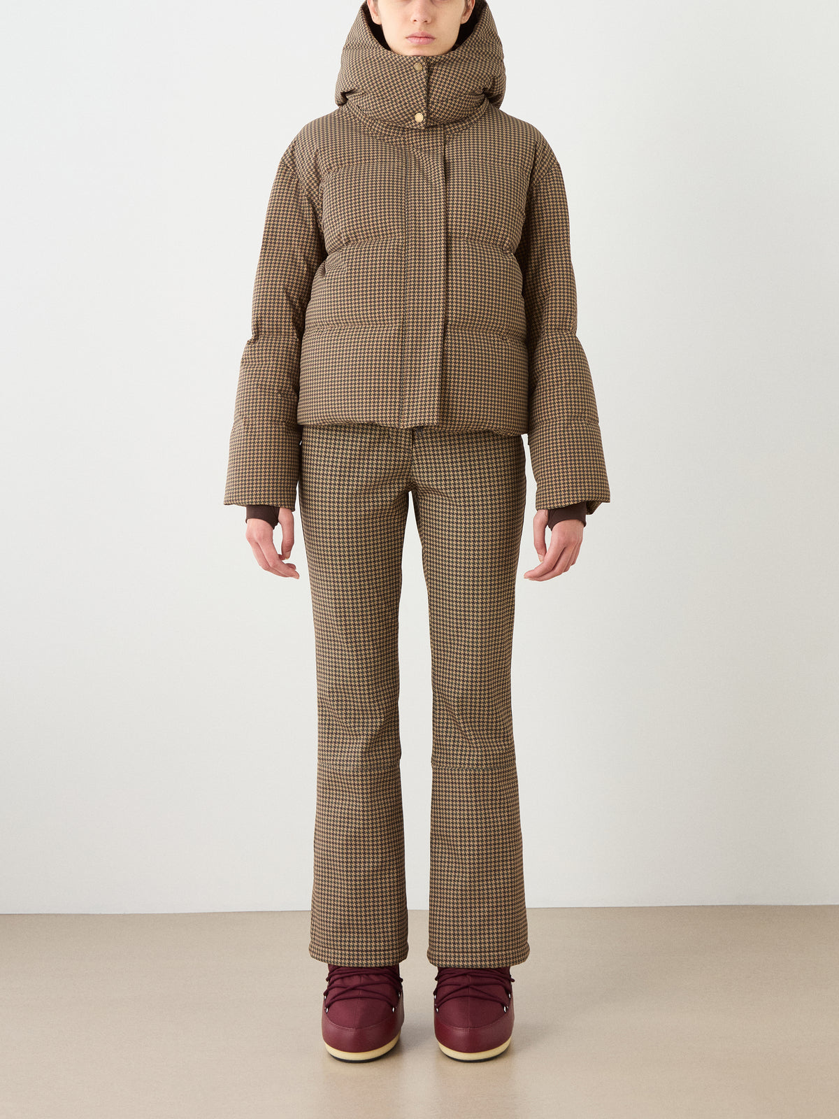 A person wears the Glenshee Ski Jacket Women by We Norwegians in brown plaid with flared pants and maroon boots, standing facing forward against a plain background.