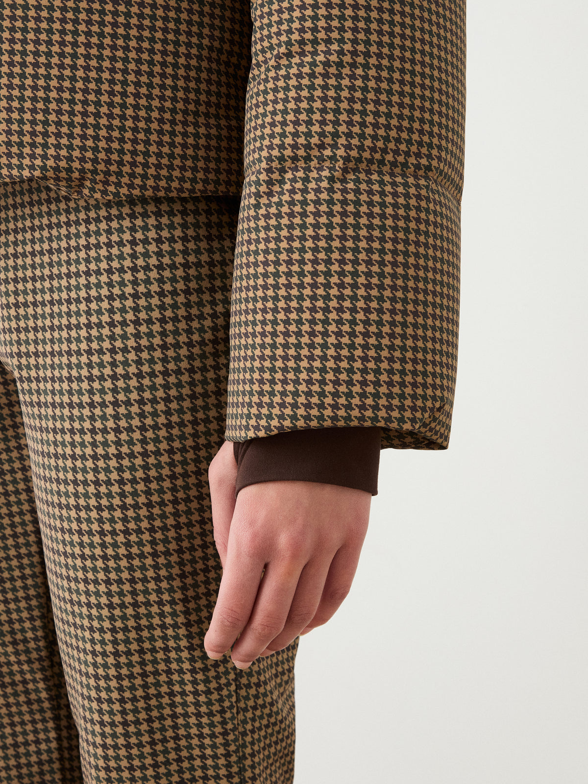 A person’s arm and hand are shown wearing the We Norwegians Glenshee Ski Jacket Women in a brown and beige houndstooth pattern, with a brown cuff at the wrist against a plain background.