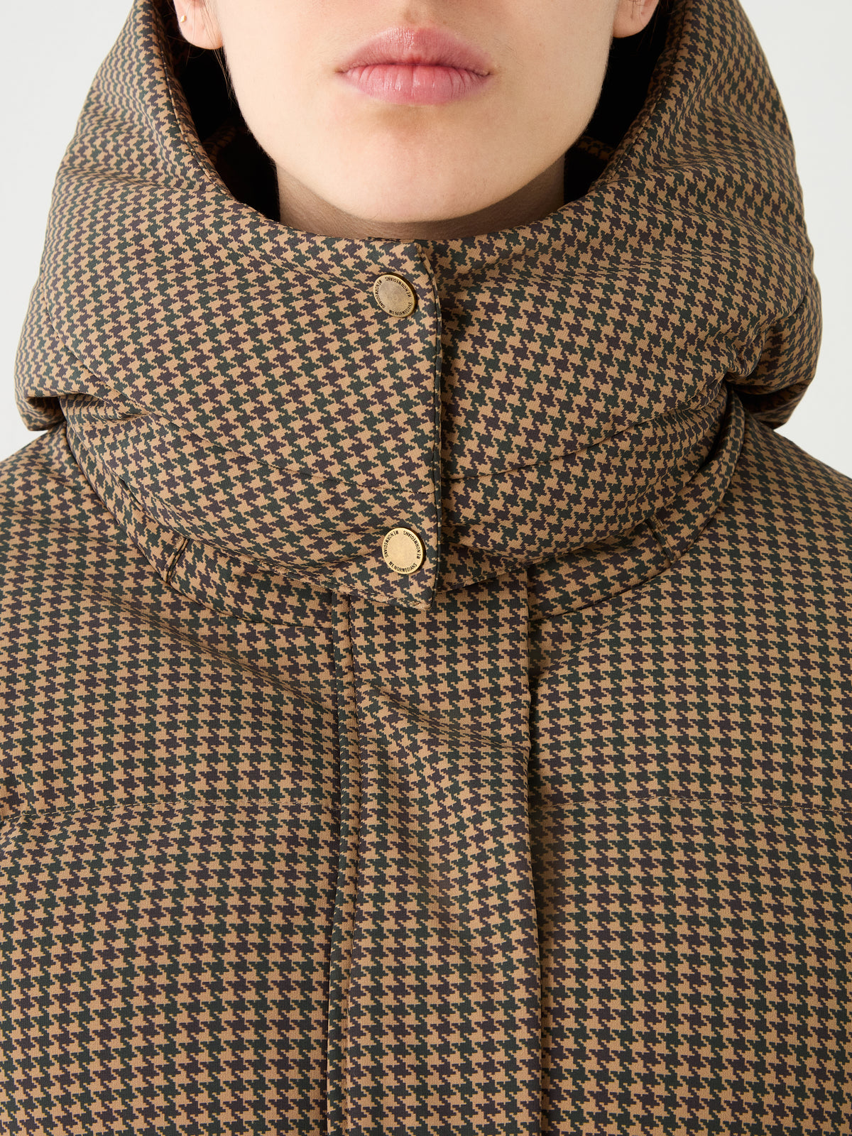 A close-up of a person wearing the Glenshee Ski Jacket Women by We Norwegians, showcasing its brown and black houndstooth pattern, high collar, and gold snap buttons. Only their lips, chin, and neck are visible.