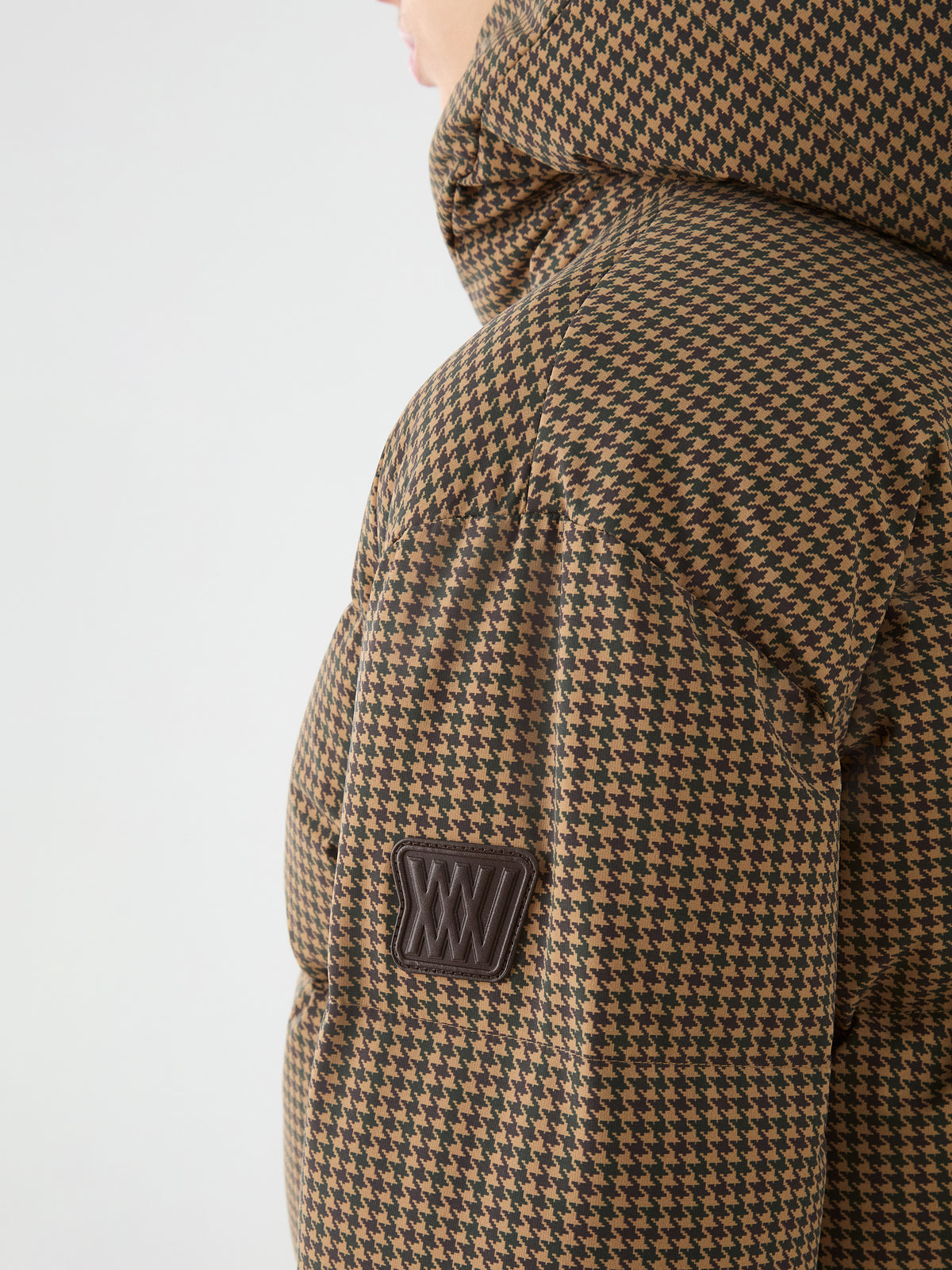 Close-up of the We Norwegians Glenshee Ski Jacket Women in brown and black houndstooth with down filling and a black sleeve patch, worn against a plain light background.
