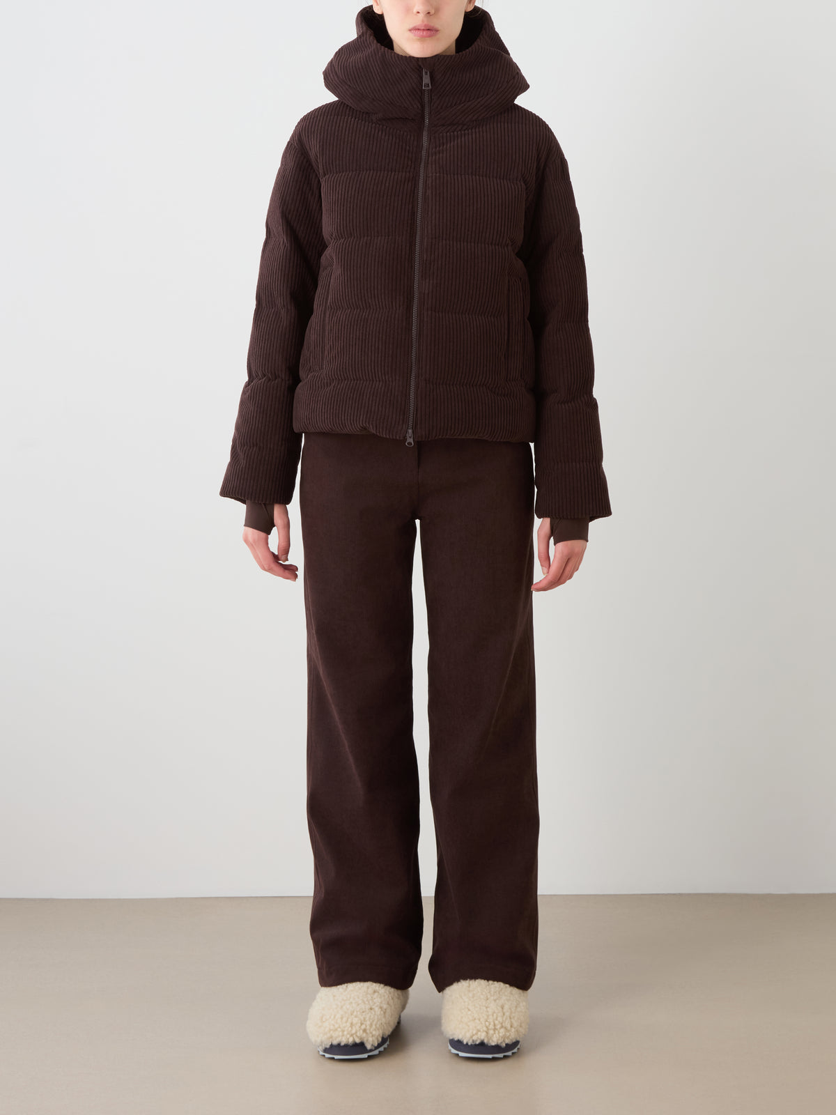 A person wears the We Norwegians Britt Ski Jacket Women in brown with matching pants and cream slippers, standing against a plain light background.