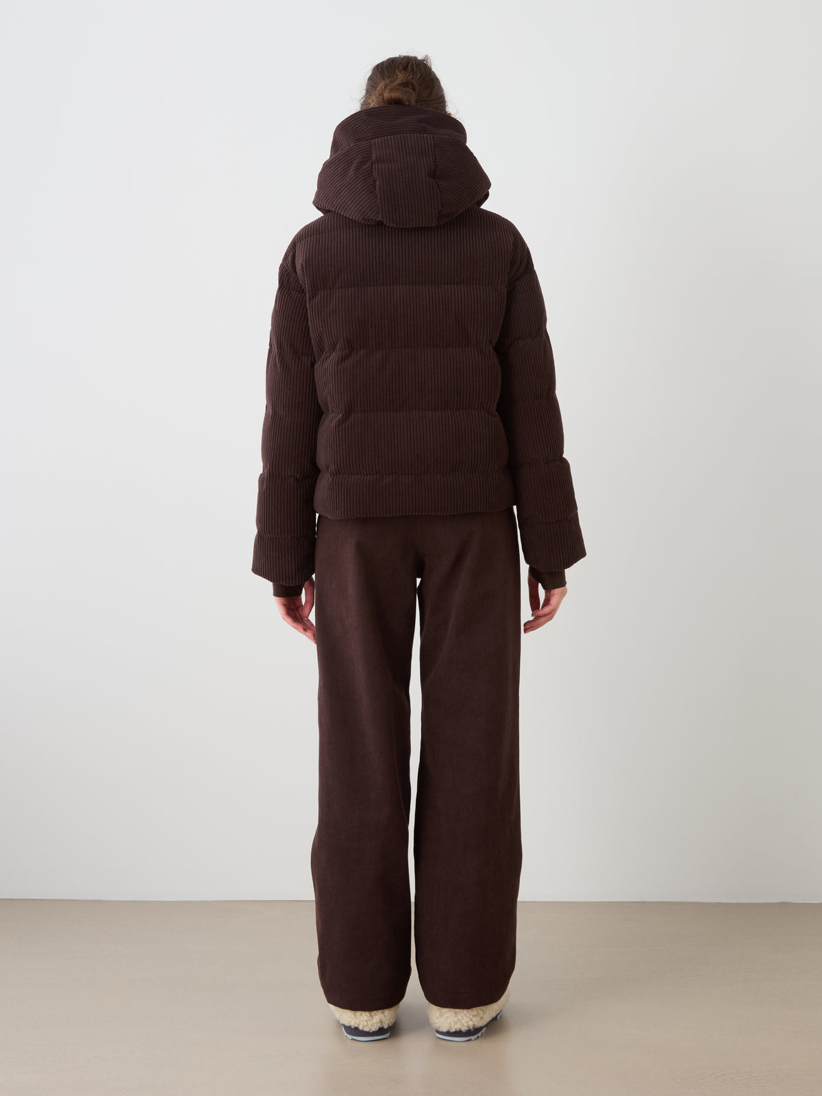 A person stands facing away, wearing the Britt Ski Jacket Women by We Norwegians in dark brown with a hood and matching wide-leg pants, on a light floor against a plain white wall.