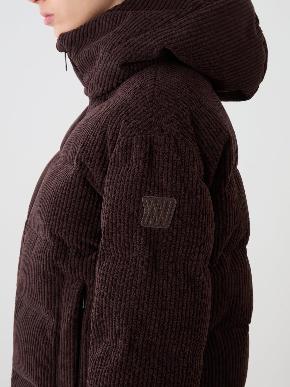 A person wears the We Norwegians Britt Ski Jacket Women in dark brown corduroy with a hood, featuring a Roman numeral XXV patch on the sleeve. The photo is cropped to show only the upper torso and part of the face.