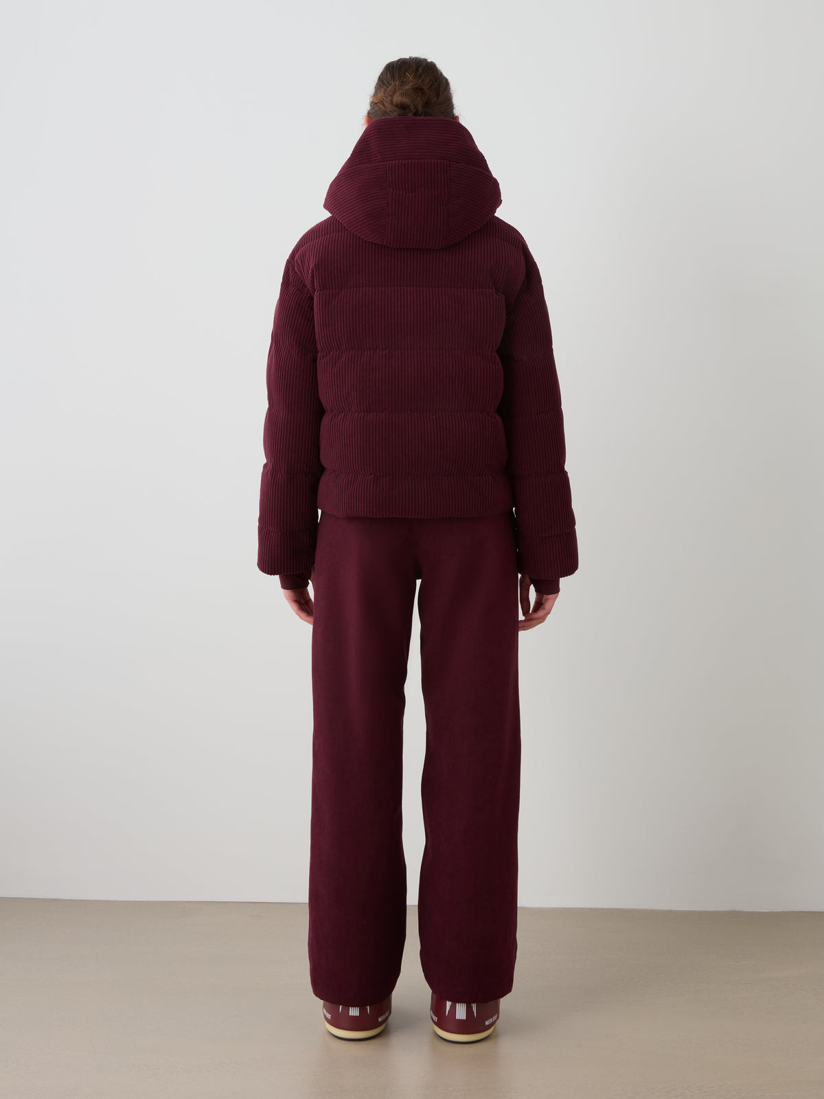 A person stands indoors, back to the camera, wearing a We Norwegians Britt Ski Jacket Women in burgundy with matching wide-leg pants and sneakers against a plain, neutral background.