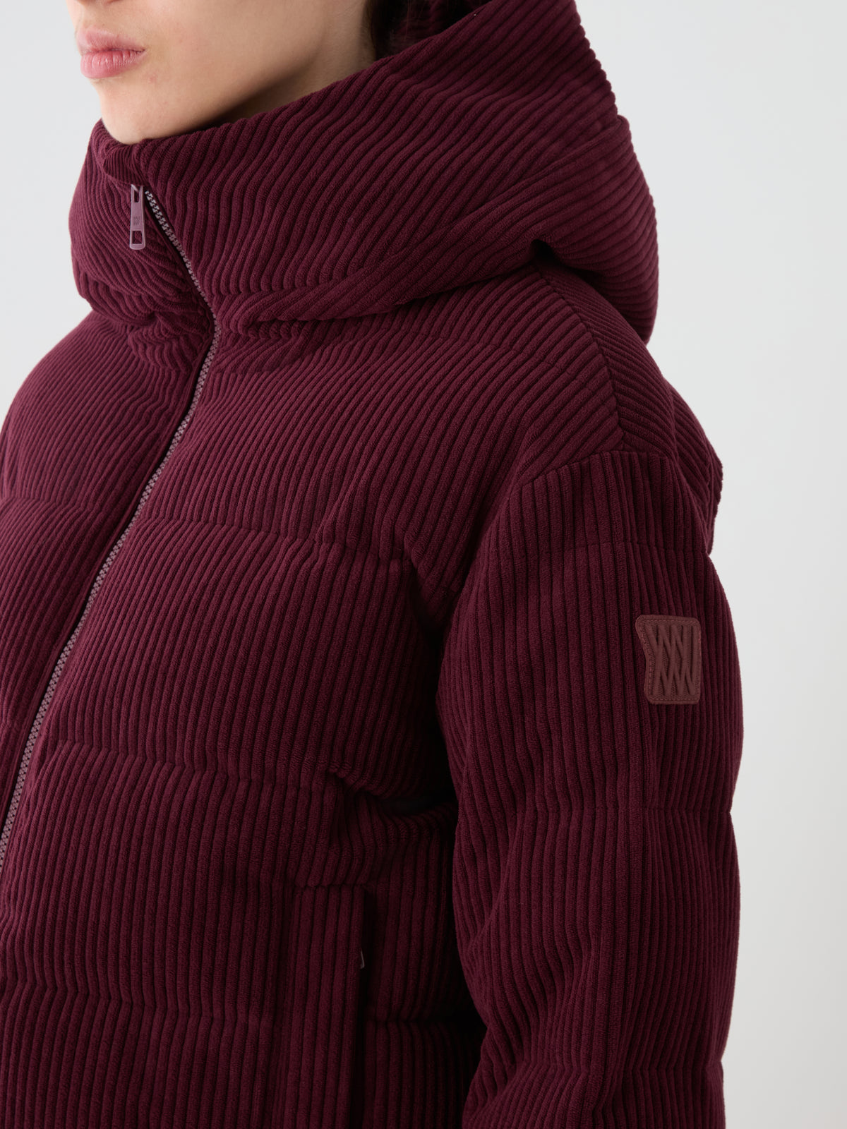 A person wears the We Norwegians Britt Ski Jacket Women in burgundy corduroy with a hood, logo patch on the sleeve, and front zipper; their face is partially out of frame.