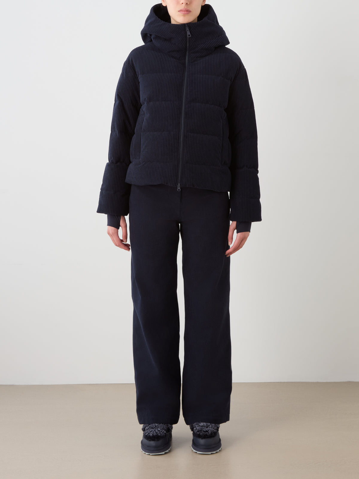 A person stands against a plain background wearing a dark We Norwegians Britt Ski Jacket Women with a hood, wide-leg pants, and dark shoes. Their face is partially hidden by the hood.