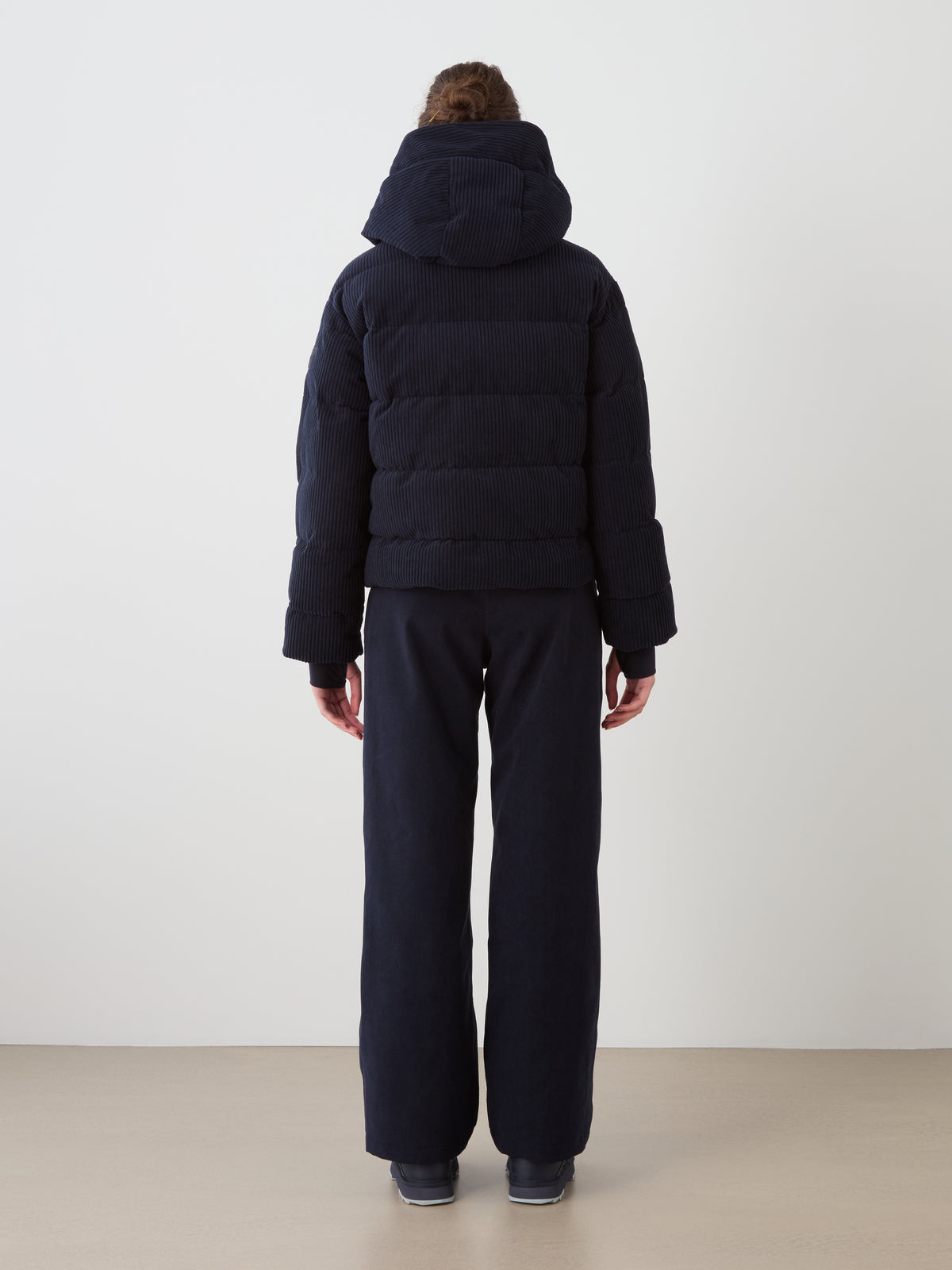 A person faces away wearing the We Norwegians Britt Ski Jacket Women in black with a hood and matching black pants. The background is plain light, and the floor is beige.