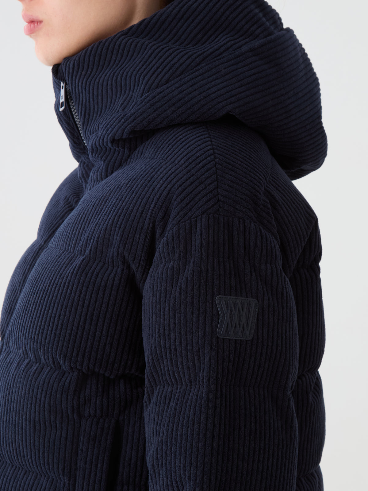 A woman in a dark blue We Norwegians Britt Ski Jacket Women with hood, seen from the side. The corduroy jacket has a raised texture and a small stitched patch on the sleeve.