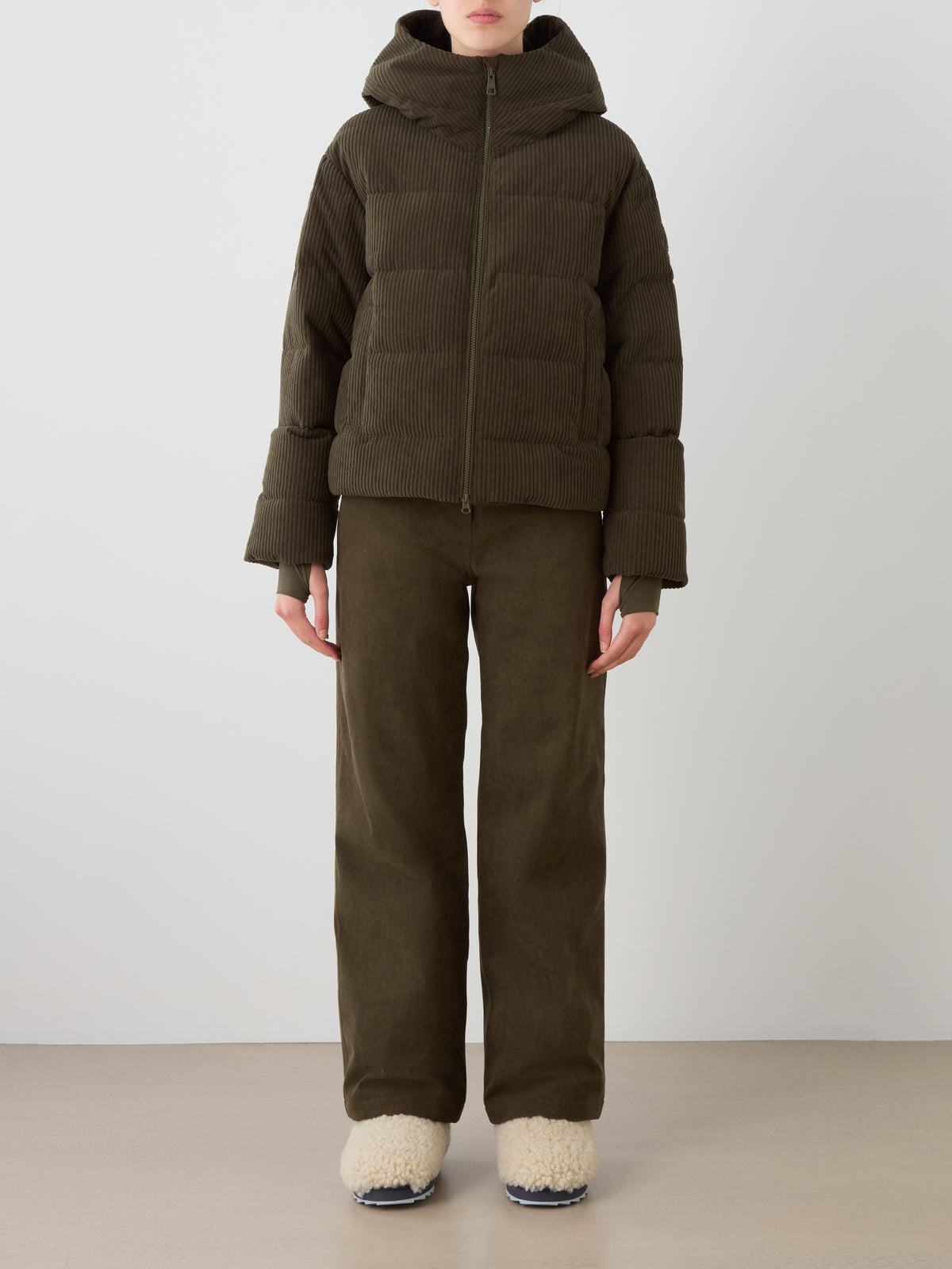 A person wears the Britt Ski Jacket Women by We Norwegians—an olive green corduroy hooded jacket paired with matching wide-leg pants and cream fuzzy slippers, styled against a plain background.