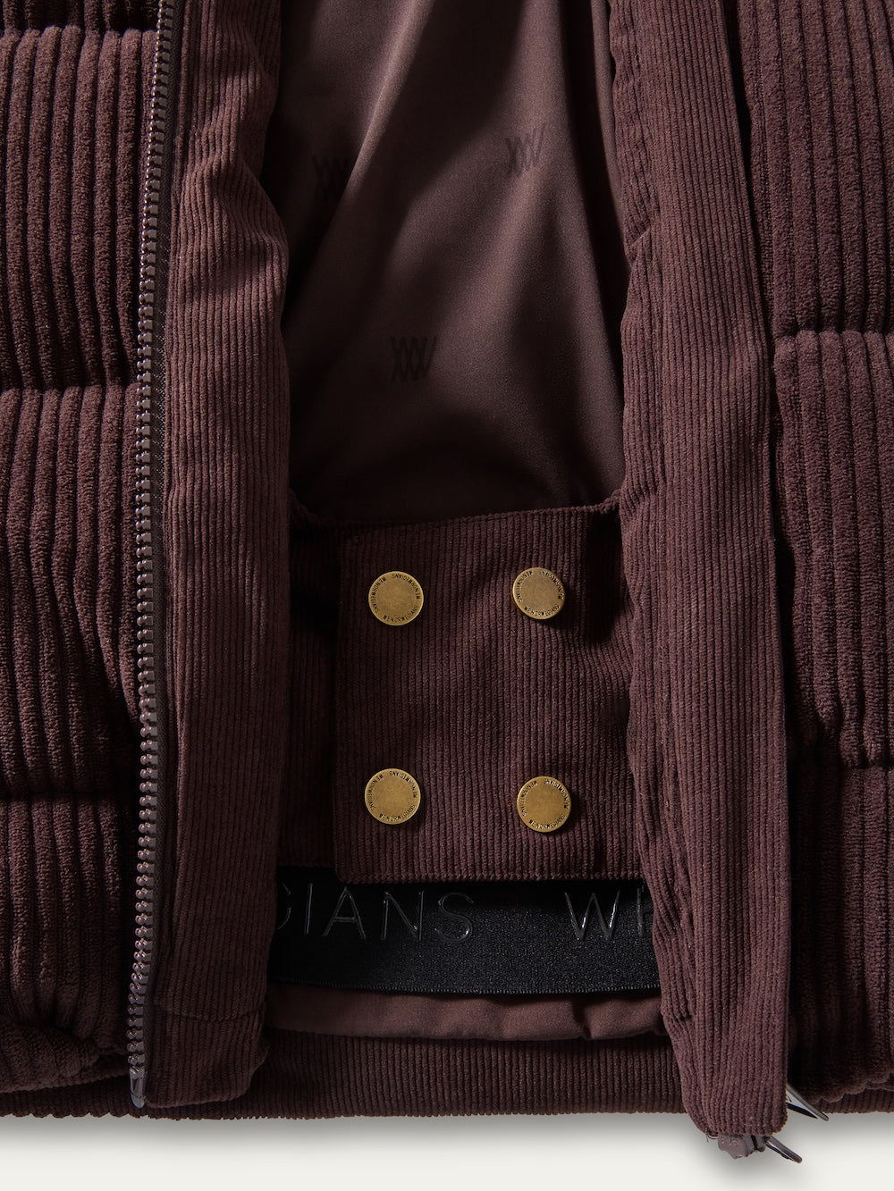 Close-up of the Britt Ski Jacket Women by We Norwegians, featuring brown corduroy, a zipper, two gold snap buttons, and an inner lining with a subtle “W” pattern plus black elastic band with white lettering.
