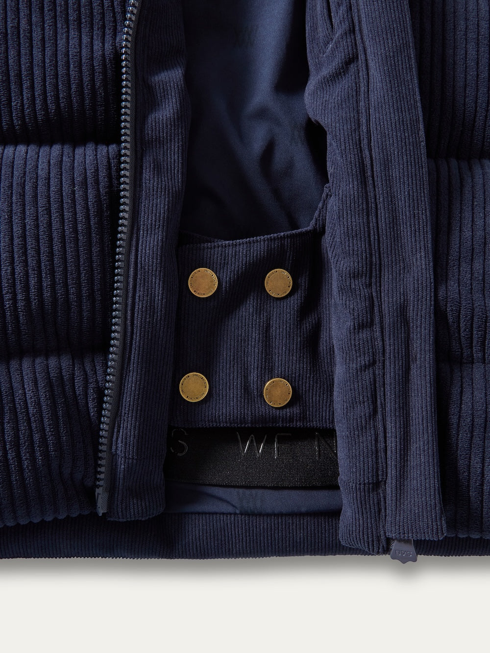 Close-up of the Britt Ski Jacket Women by We Norwegians, a navy blue corduroy ski jacket with gold snap buttons and a black elastic hem, shown partially unzipped to reveal the inner lining.