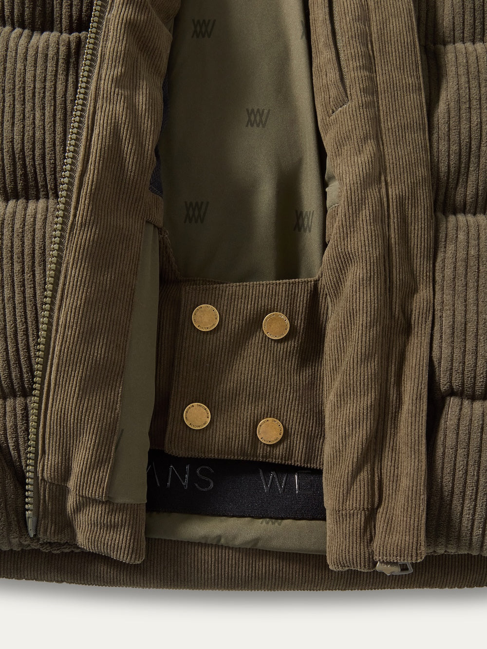 Close-up of the We Norwegians Britt Ski Jacket Women in olive green corduroy, partially unzipped to reveal its “X” patterned lining and snap button placket with three brass buttons.