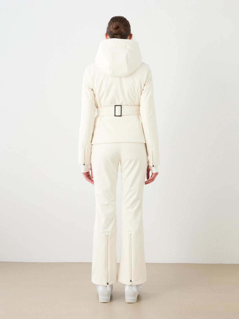 A person stands facing away in the We Norwegians Tyin Tailored Ski Jacket Women with a belt and matching pants, plus white boots, against a plain light background.