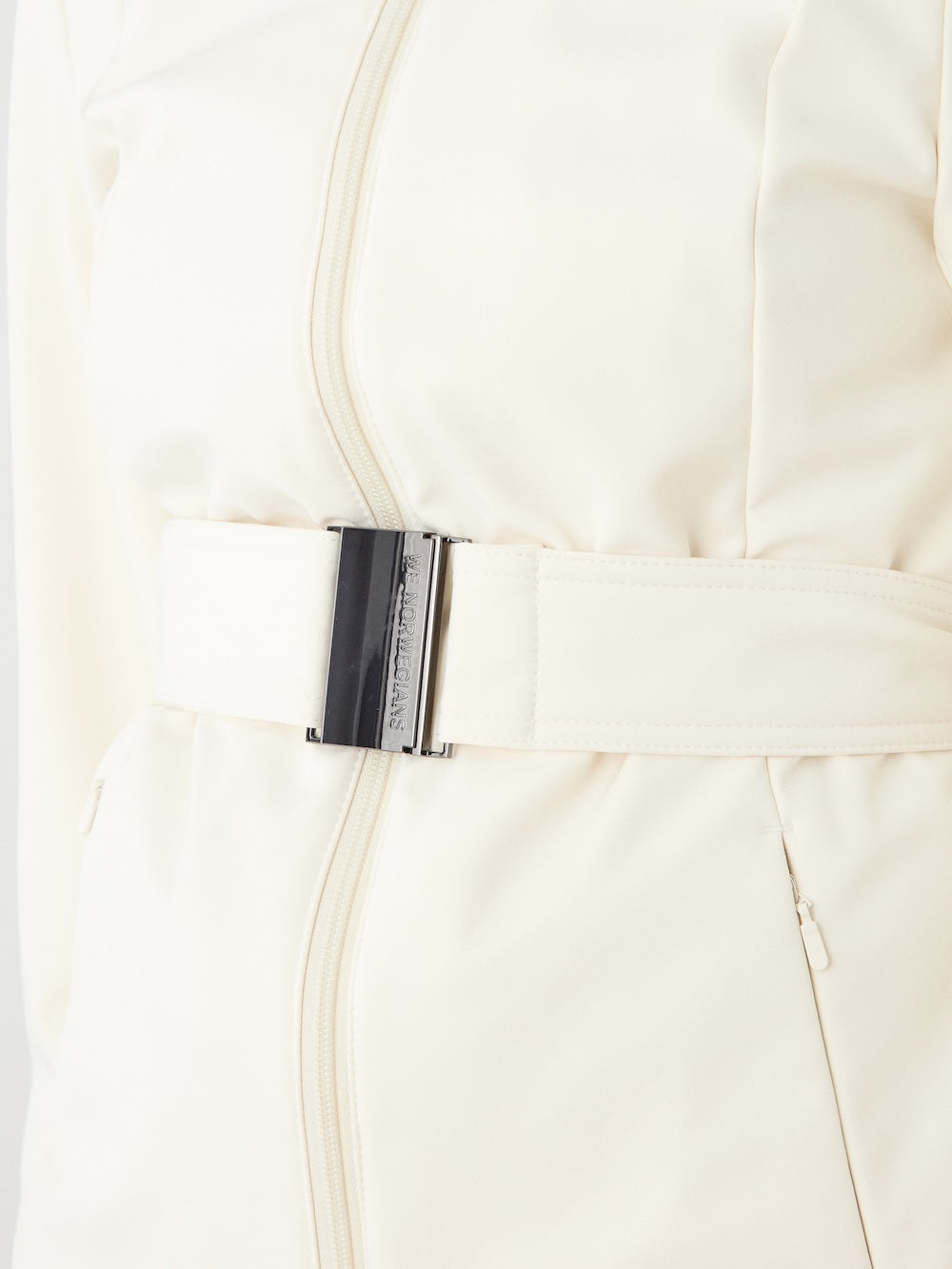 Close-up of the We Norwegians Tyin Tailored Ski Jacket Women in cream, featuring a matching belt with a metallic rectangular buckle and a vertical zipper beneath the belt.