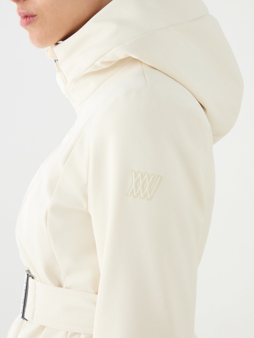 A person wears the We Norwegians Tyin Tailored Ski Jacket Women in off-white, featuring a belted waist and “XXXV” embroidery on the upper sleeve, with their lower face partially visible.