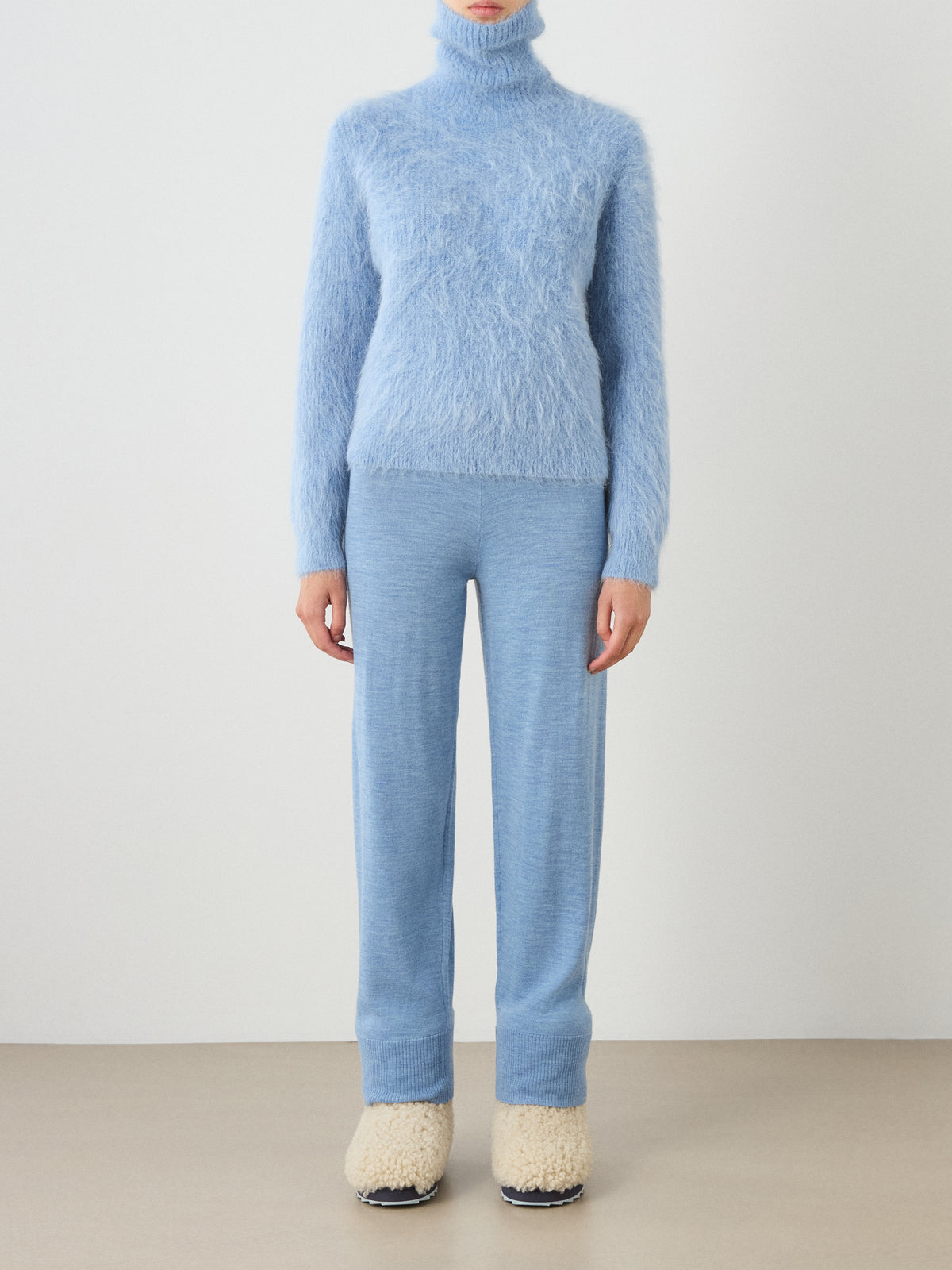 A person wearing the We Norwegians Myrull Sweater Women in a fuzzy light blue, paired with matching light blue pants and cream-colored fuzzy slippers, standing against a plain light background.