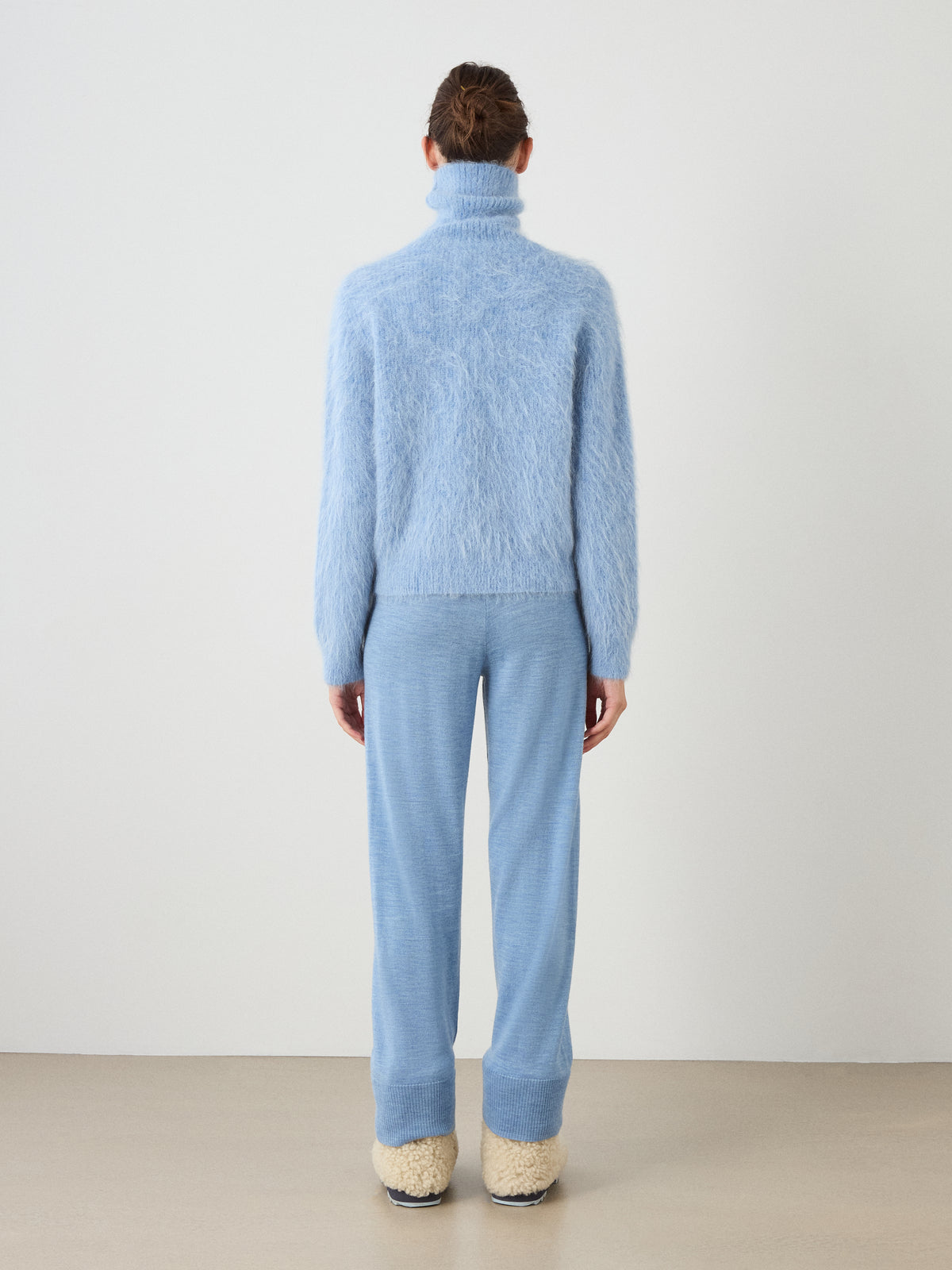 A person stands facing a white wall, wearing the We Norwegians Myrull Sweater Women in a fuzzy light blue, paired with matching light blue pants and cream-colored slippers. Their hair is tied back.