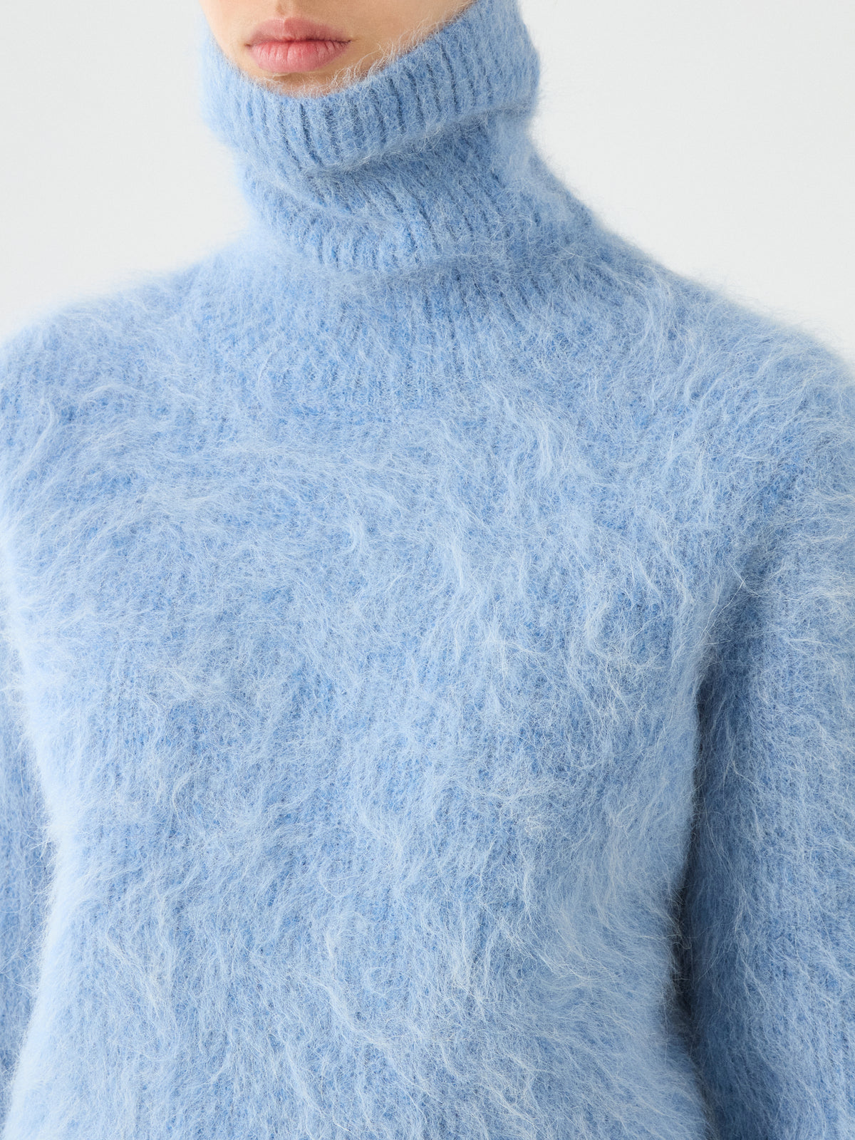 A person wearing the We Norwegians Myrull Sweater Women—a soft, fuzzy light blue alpaca turtleneck. Only the lower half of their face and upper body are visible against a plain background.