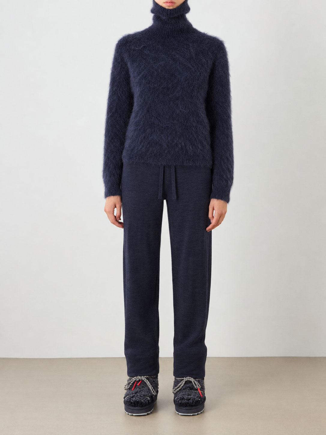 A person stands against a plain background wearing the Myrull Sweater Women by We Norwegians, a cozy navy blue alpaca turtleneck, paired with navy drawstring lounge pants and black textured slip-on shoes. Their face is not visible.