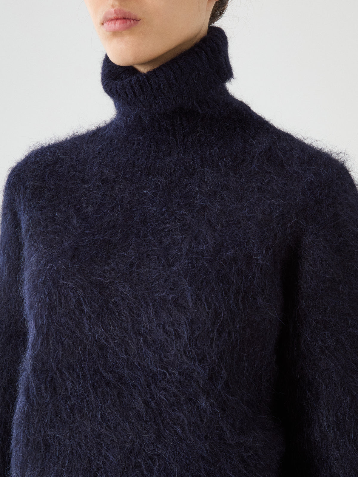 A person wearing the We Norwegians Myrull Sweater Women in fuzzy dark blue, shown from the shoulders up against a plain light background. Their face is partially visible, and their hair is pulled back.