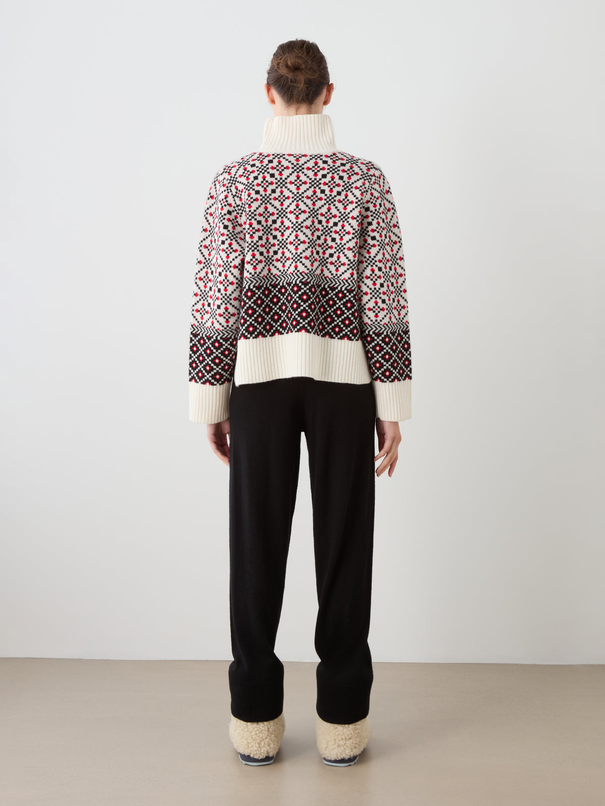 A person stands facing away, wearing the We Norwegians Telemark Sweater Women—a thick knit merino wool sweater featuring a red, white, and black pattern inspired by classic Norwegian designs—paired with black pants and cream-colored, fuzzy footwear. The background is plain and neutral.
