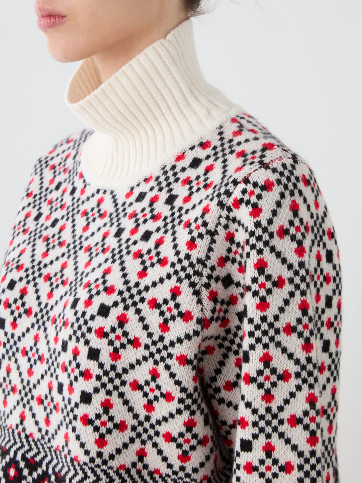 A woman is wearing the We Norwegians Telemark Sweater Women, featuring red, black, and white geometric designs inspired by classic Norwegian knitting patterns, layered over a white ribbed turtleneck. The image captures her upper torso and part of her face in profile.