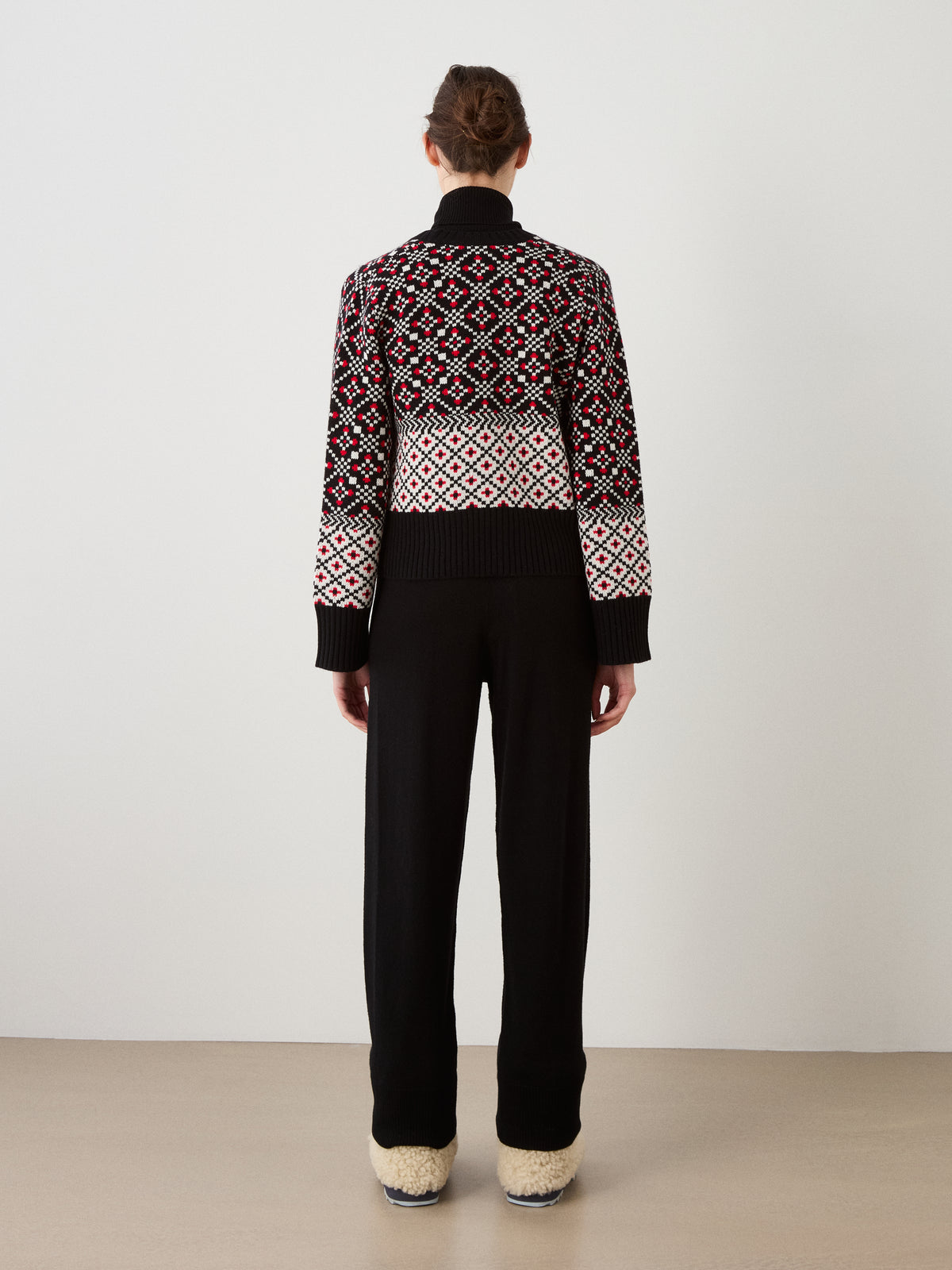A person stands facing away, wearing the We Norwegians Telemark Cardigan Women in a bold black, white, and red Nordic-inspired pattern, paired with black pants and cream-colored fuzzy shoes, set against a plain light background.
