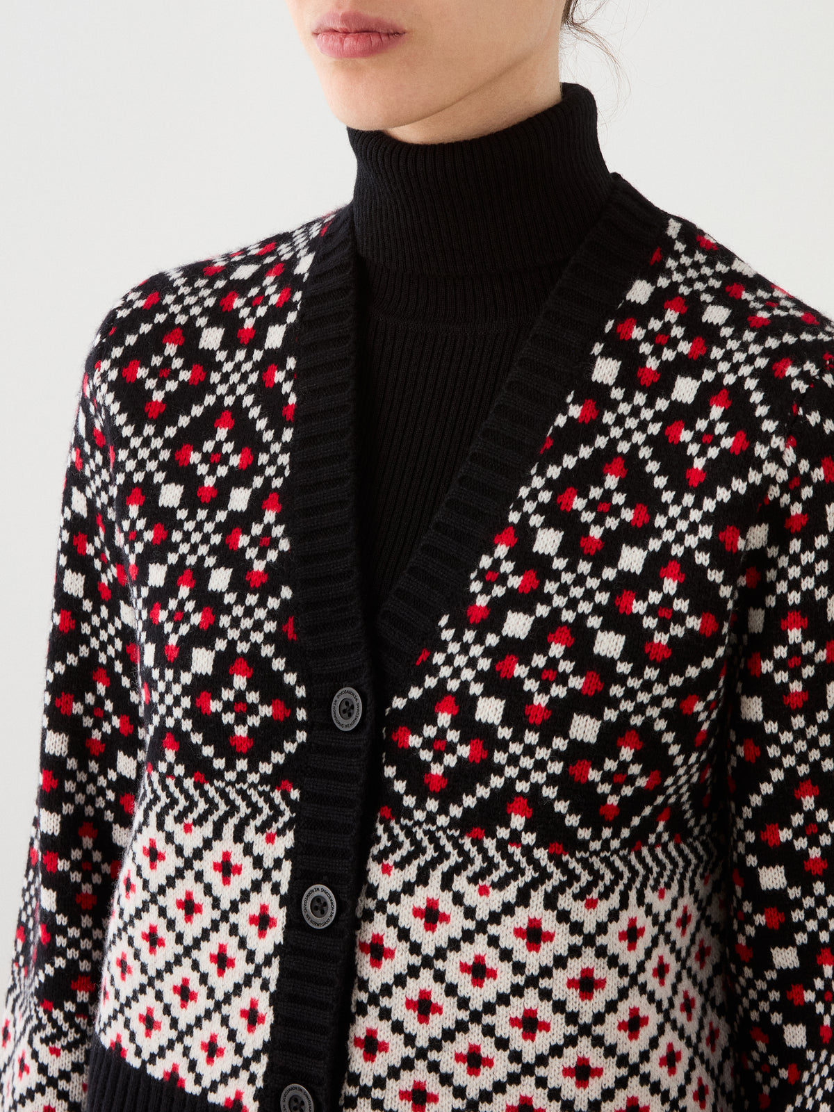 A person wearing a black turtleneck and the We Norwegians Telemark Cardigan Women, which features a red, white, and black geometric pattern inspired by classic Nordic knitwear. Only the lower face and upper torso are visible.