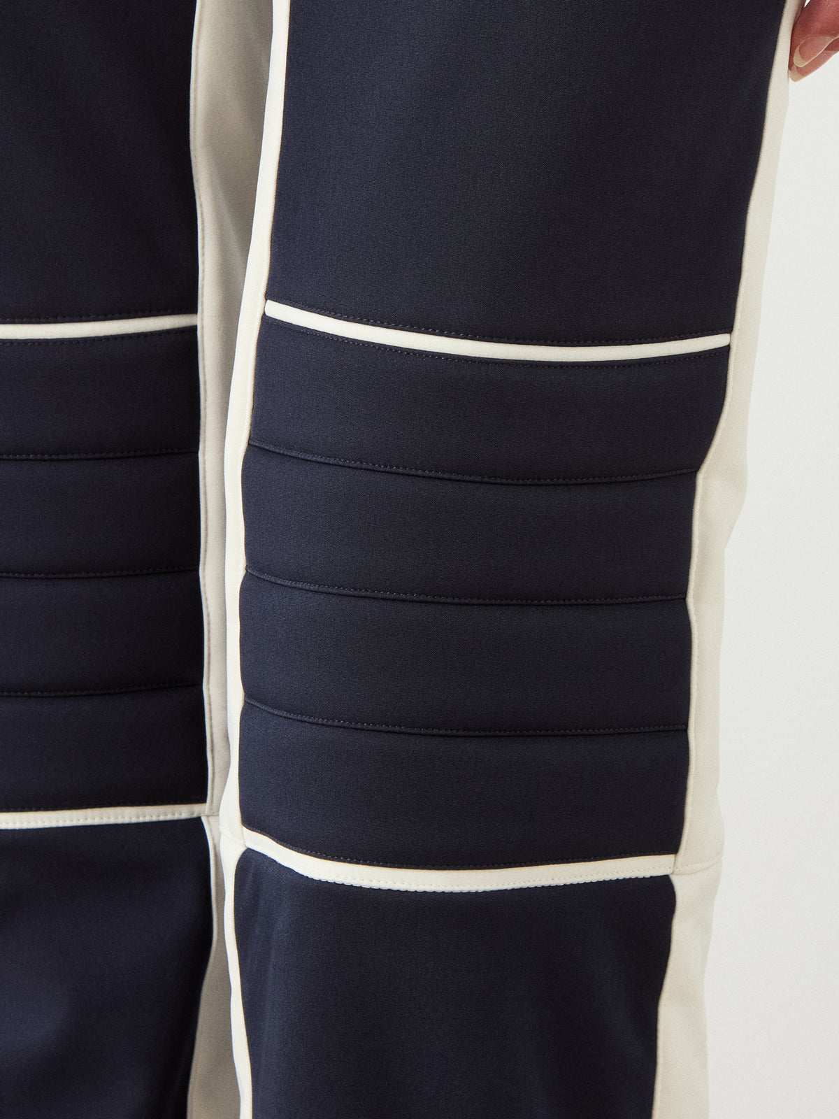 Close-up of the We Norwegians Solo Bib Pant Women shows dark blue fabric, horizontal padded sections, white side panels, and detailed stitching for a vintage alpine ski style.