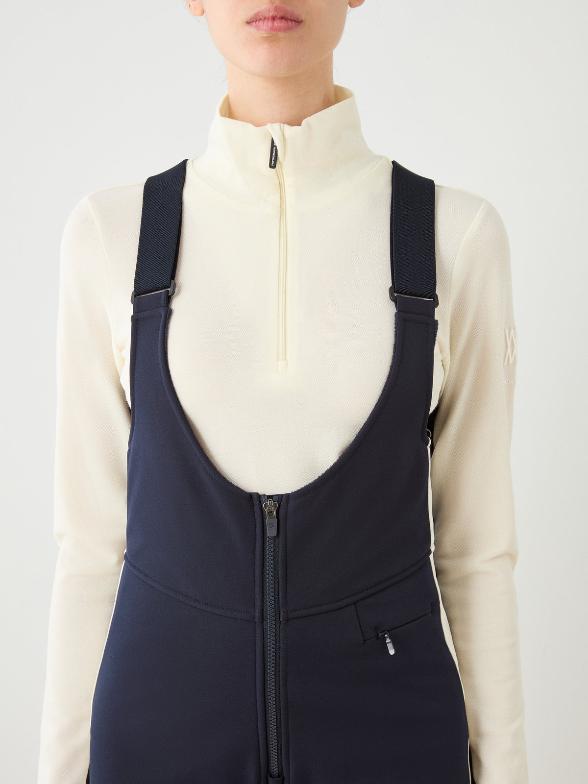 A person wearing We Norwegians Solo Bib Pant Women in navy with a front zipper, styled with a cream zip-up turtleneck, is shown from shoulders to waist against a plain white background, evoking vintage alpine ski style.