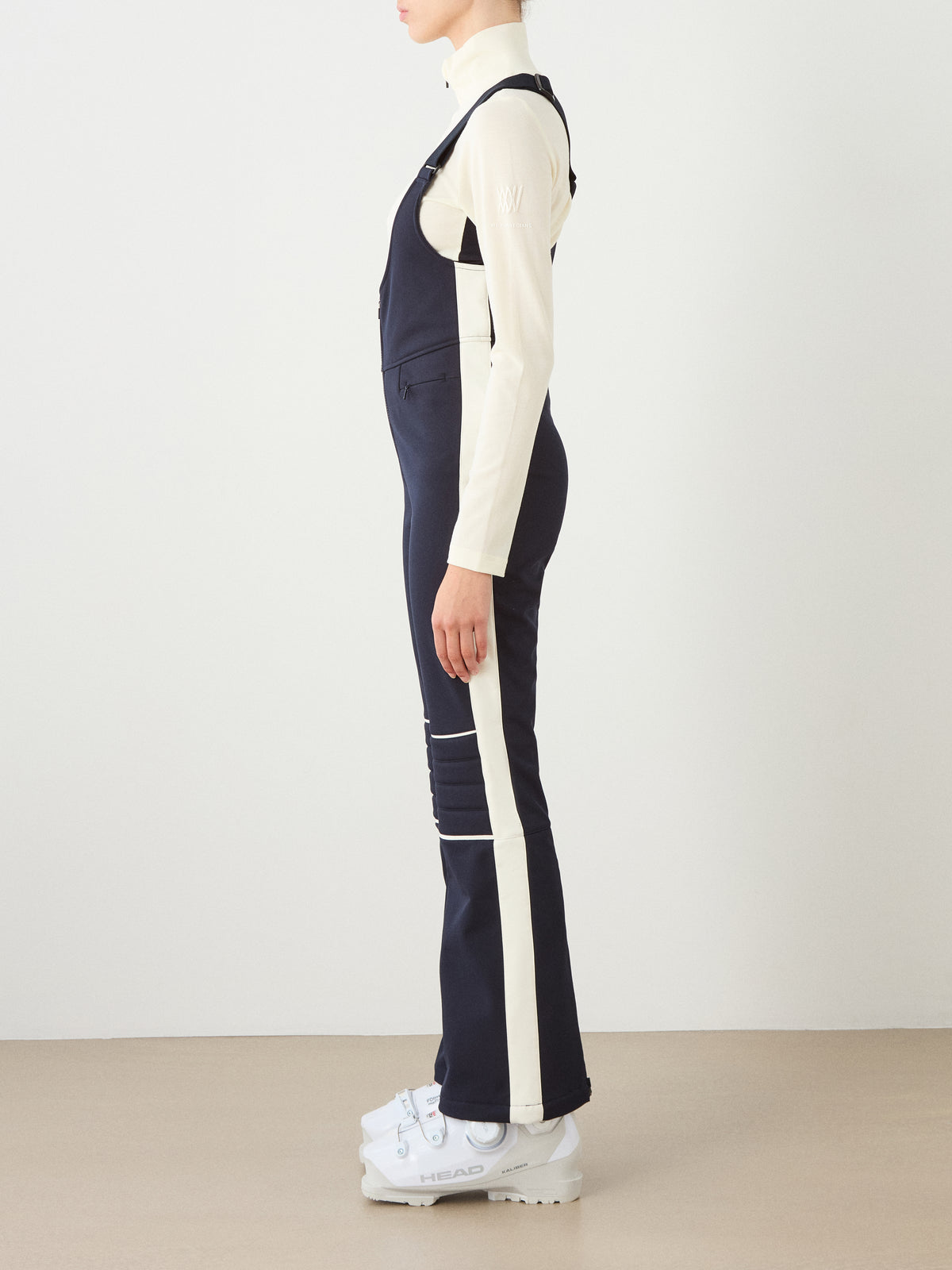 A person stands sideways wearing a cream top, We Norwegians Solo Bib Pant Women in navy and white, and white ski boots, set against a plain light background.
