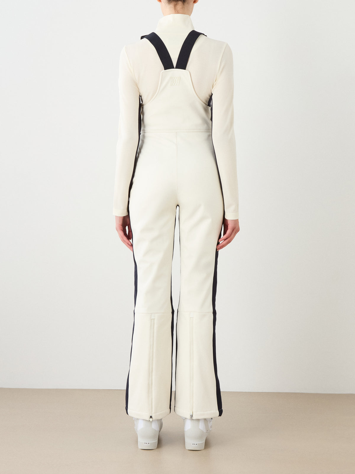 A person stands facing away from the camera, wearing the We Norwegians Solo Bib Pant Women—white with black side stripes, wide straps—and white boots against a plain background.