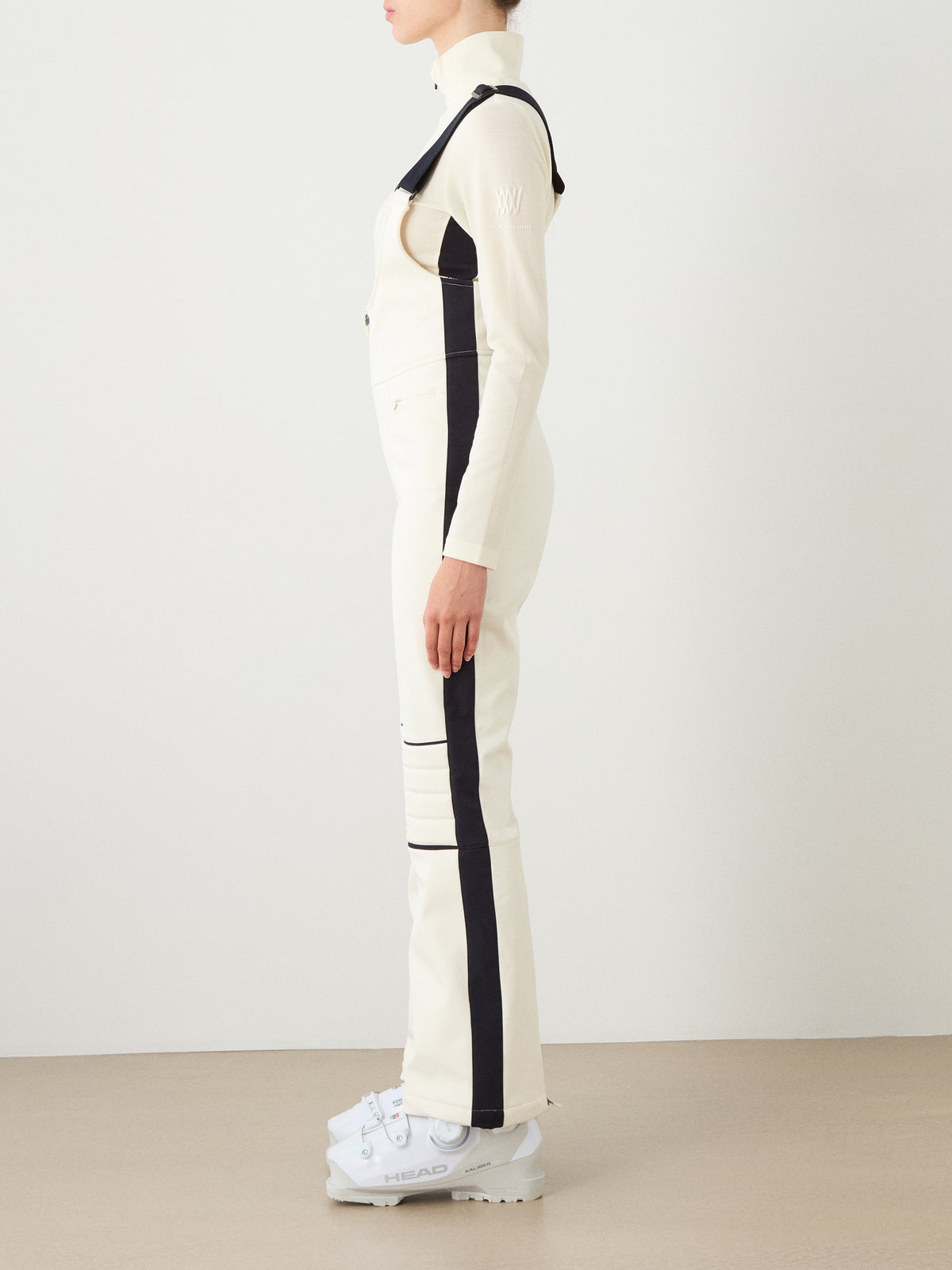 A person stands sideways in the cream-colored We Norwegians Solo Bib Pant Women with black stripes and white ski boots, evoking vintage alpine style against a plain light background.