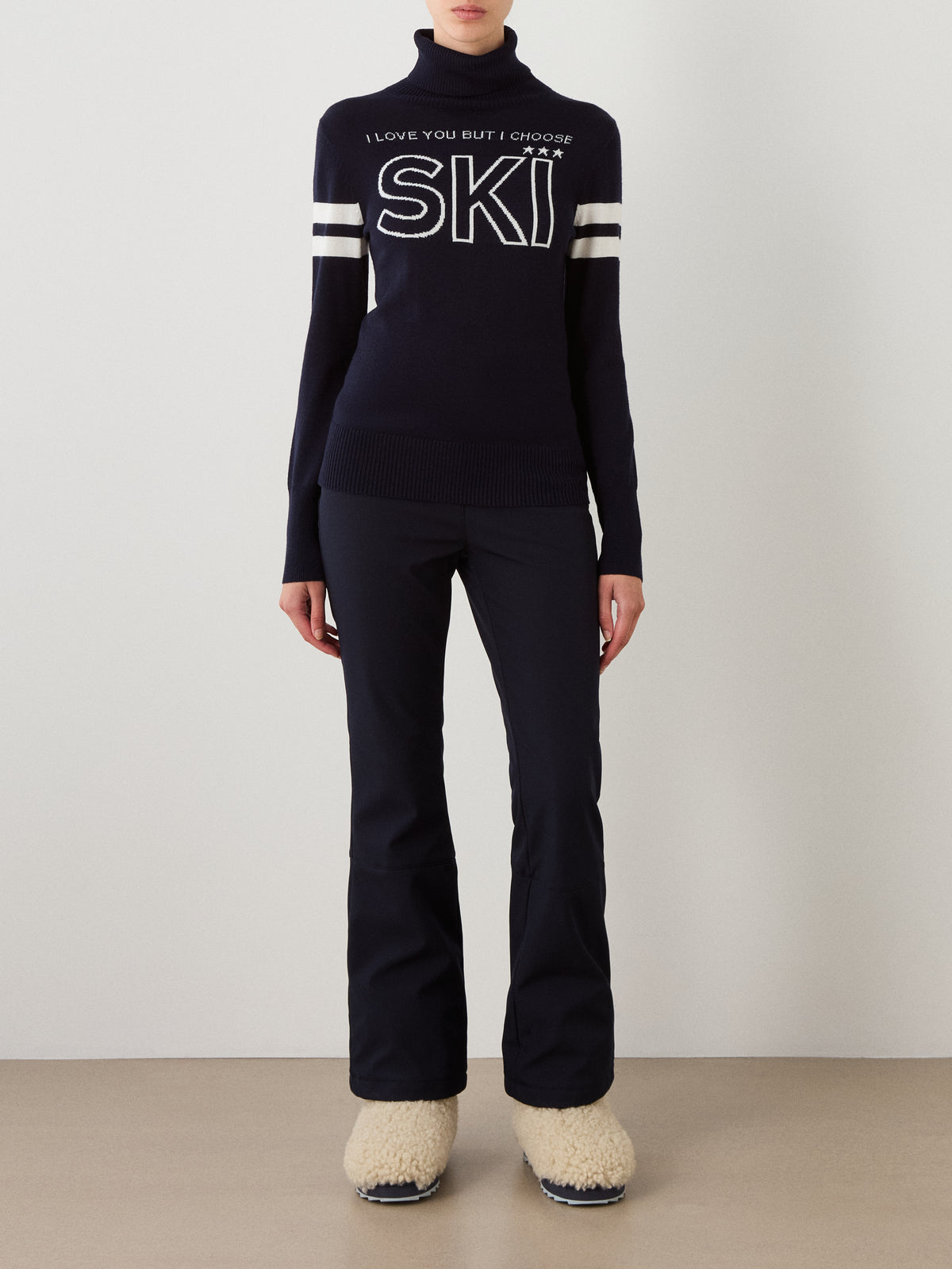 A person stands indoors against a plain white wall and tan floor, channeling retro ski fashion in the We Norwegians Bibi Fitted Sweater Women—a navy merino wool sweater featuring white stripes and “I LOVE YOU BUT I CHOOSE SKI” text—paired with navy pants and beige fuzzy boots.
