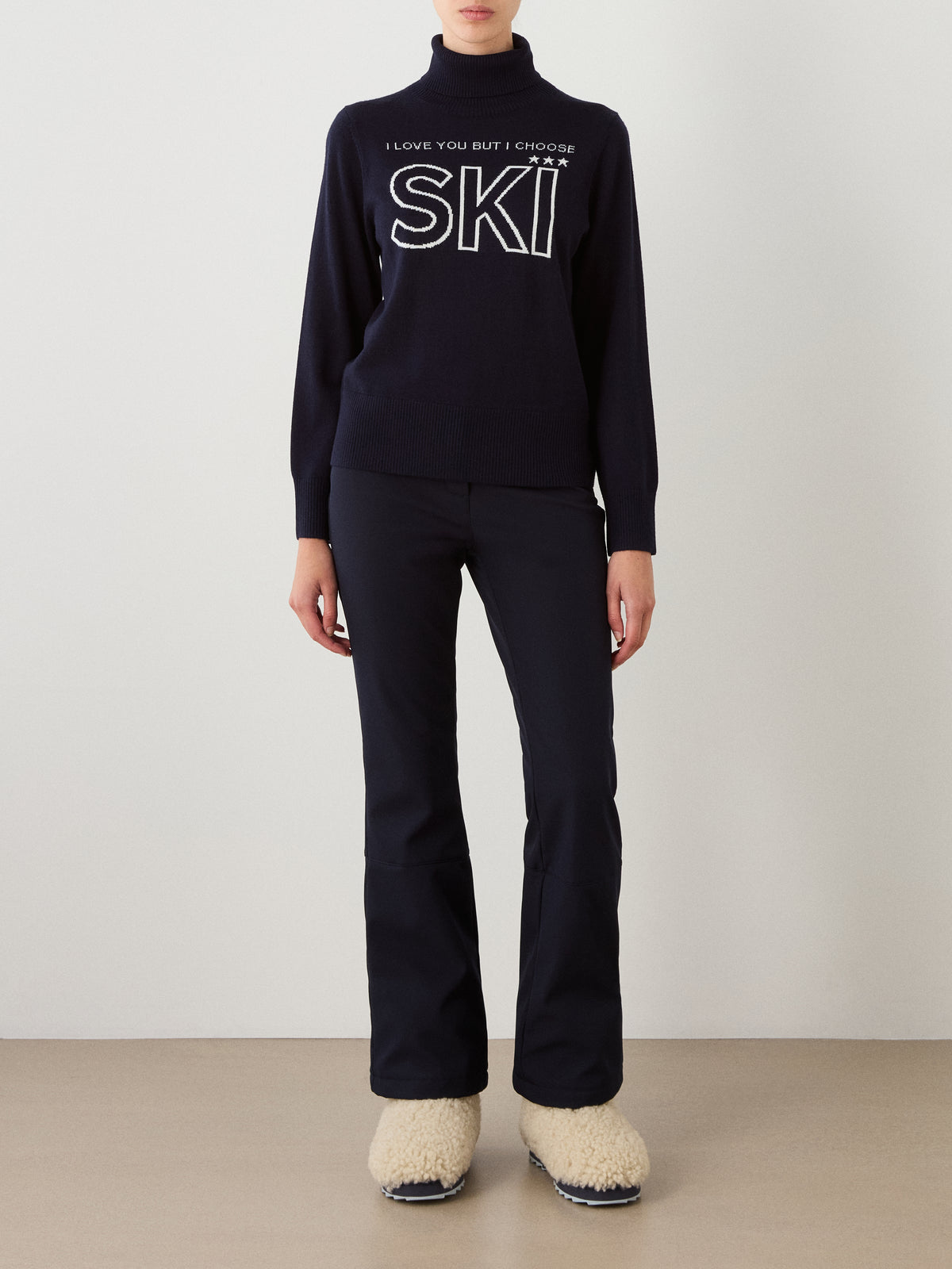 A person wearing the We Norwegians Bibi Sweater Women in black, featuring the “I LOVE YOU BUT I CHOOSE SKI” text, paired with black pants and cream, fluffy shoes, stands against a plain light background—a perfect tribute to retro ski style.