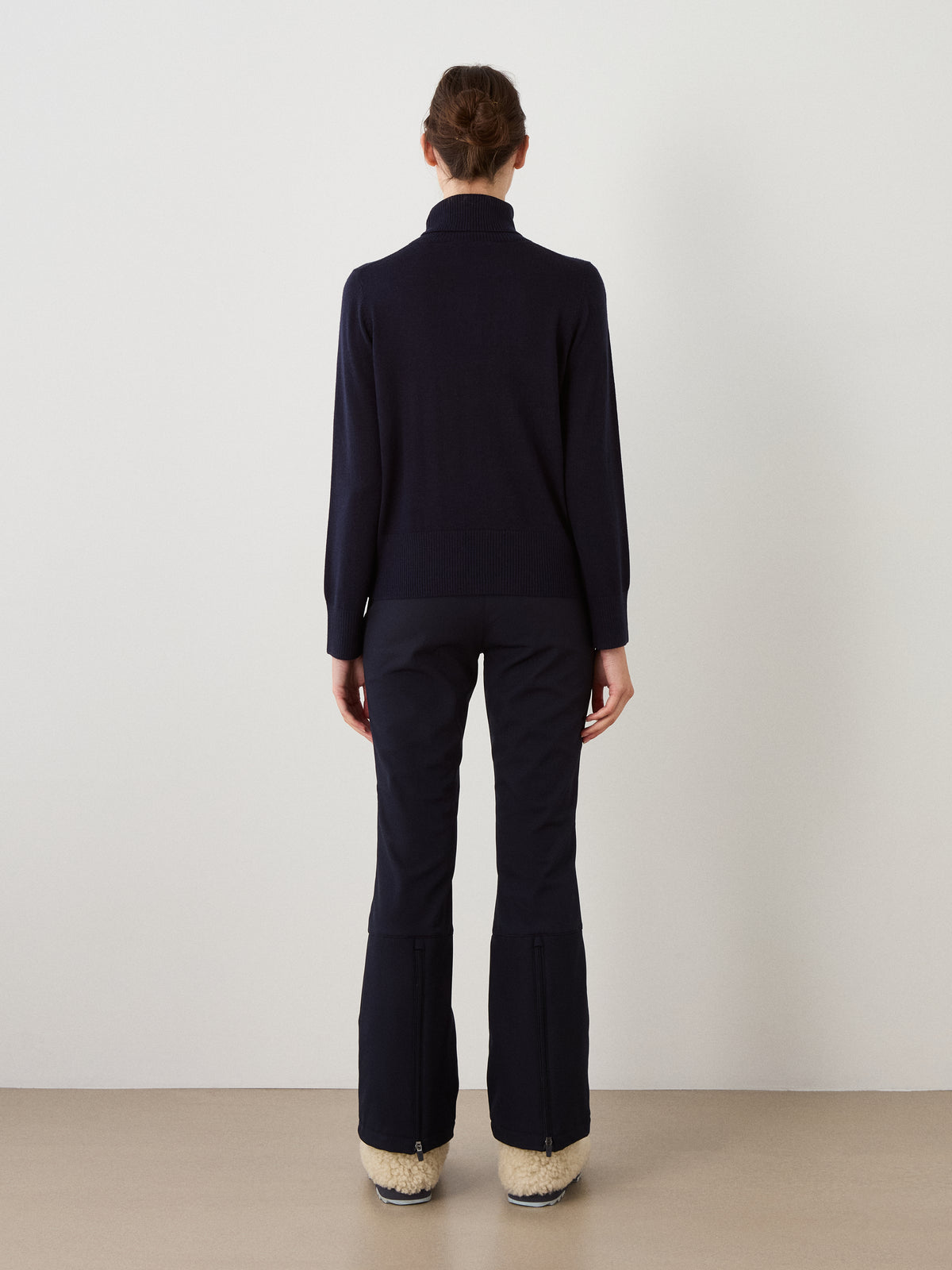 A person faces a white wall, wearing the We Norwegians Bibi Sweater Women in a dark long-sleeve style with matching dark pants and beige winter boots. The retro ski-inspired look is highlighted by the cozy sweater and relaxed stance, viewed from behind as their arms rest naturally at their sides.