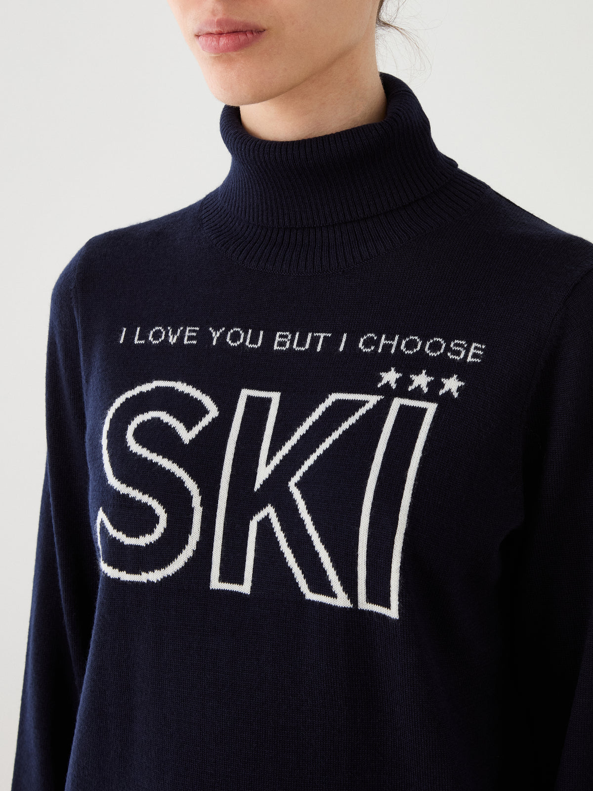 A person wearing the We Norwegians Bibi Sweater Women in navy, styled in retro ski fashion, with the words “I LOVE YOU BUT I CHOOSE SKI” and three stars above large “SKI” lettering on the front.