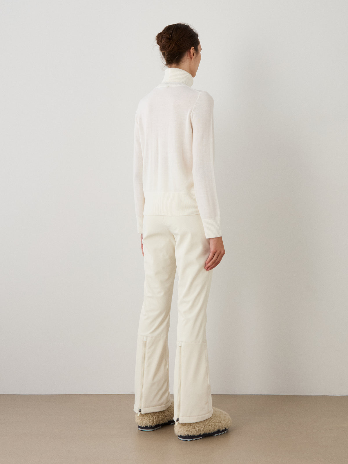 A person with brown hair, dressed in an all-white ensemble and fuzzy beige boots, stands indoors facing a white wall with their back to the camera. Their outfit is topped with the We Norwegians Bibi Sweater Women, a piece inspired by retro ski fashion that adds a chic yet nostalgic vibe to the look.
