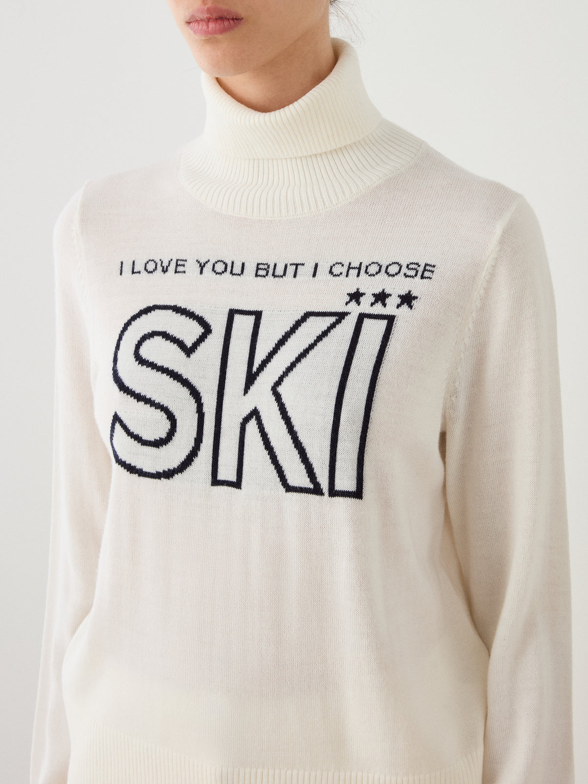 A woman wearing the We Norwegians Bibi Sweater Women—a white merino wool ski sweater in retro ski fashion, featuring the phrase “I LOVE YOU BUT I CHOOSE SKI” and three stars above “SKI”—makes a perfect statement piece for fans of the iconic Bibi Sweater style.