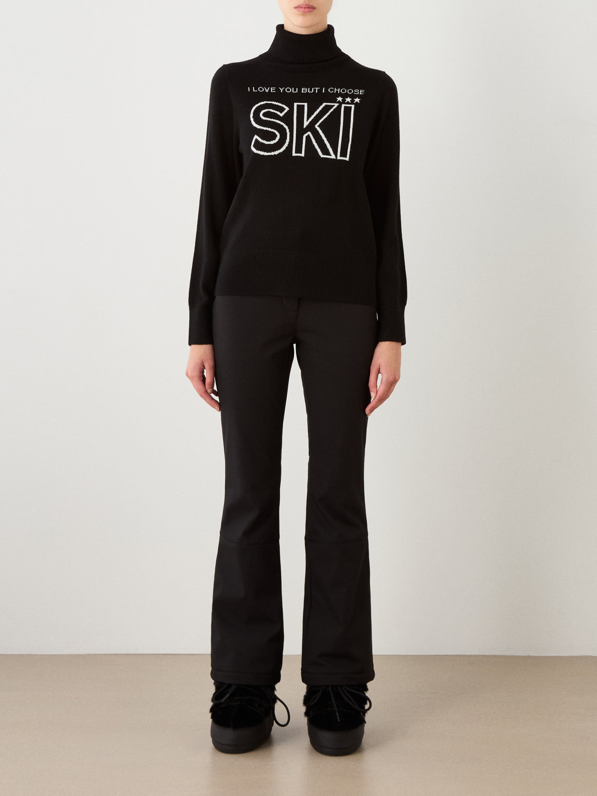 A person stands against a plain background, wearing the We Norwegians Bibi Sweater Women—a black merino wool ski sweater featuring the phrase “I LOVE YOU BUT I CHOOSE SKI”—paired with black flared pants and black footwear, channeling retro ski fashion vibes.