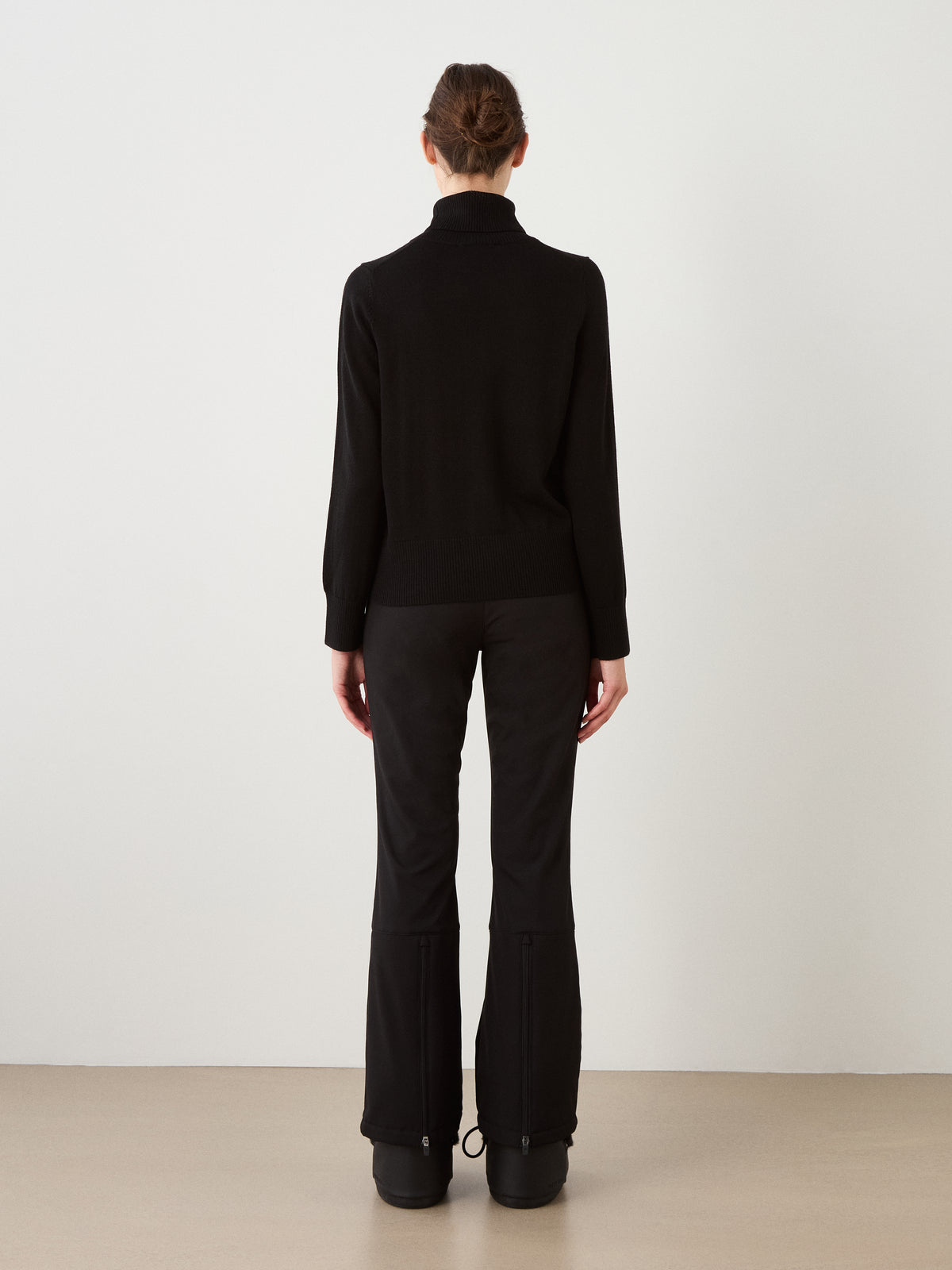 A woman stands facing away from the camera, wearing the We Norwegians Bibi Sweater in black, paired with black pants and black shoes, channeling retro ski style against a plain white background.