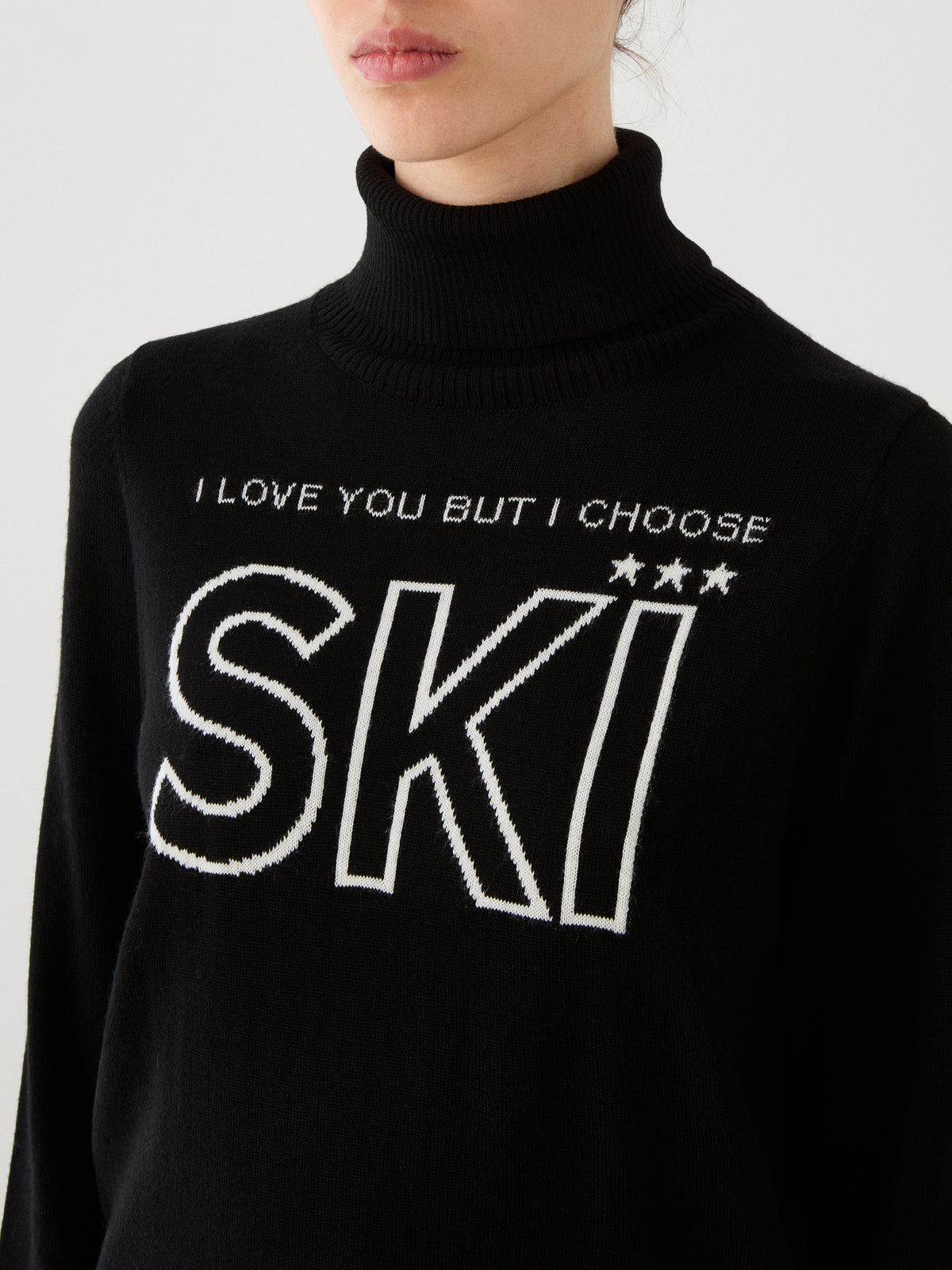 A woman dressed in the Bibi Sweater Women by We Norwegians—a black turtleneck featuring “I LOVE YOU BUT I CHOOSE SKI” and three white stars on the front—captures the essence of retro ski style.