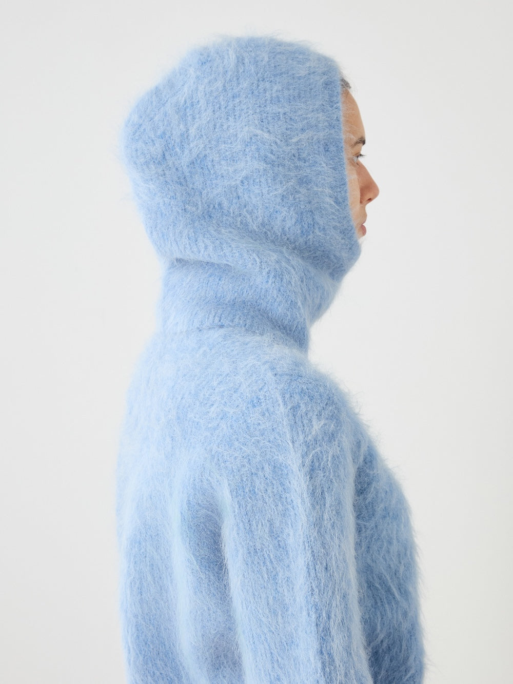 A person in profile wears the Myrull Balaclava by We Norwegians, a light blue fuzzy alpaca hooded sweater with the hood up, set against a plain white background.