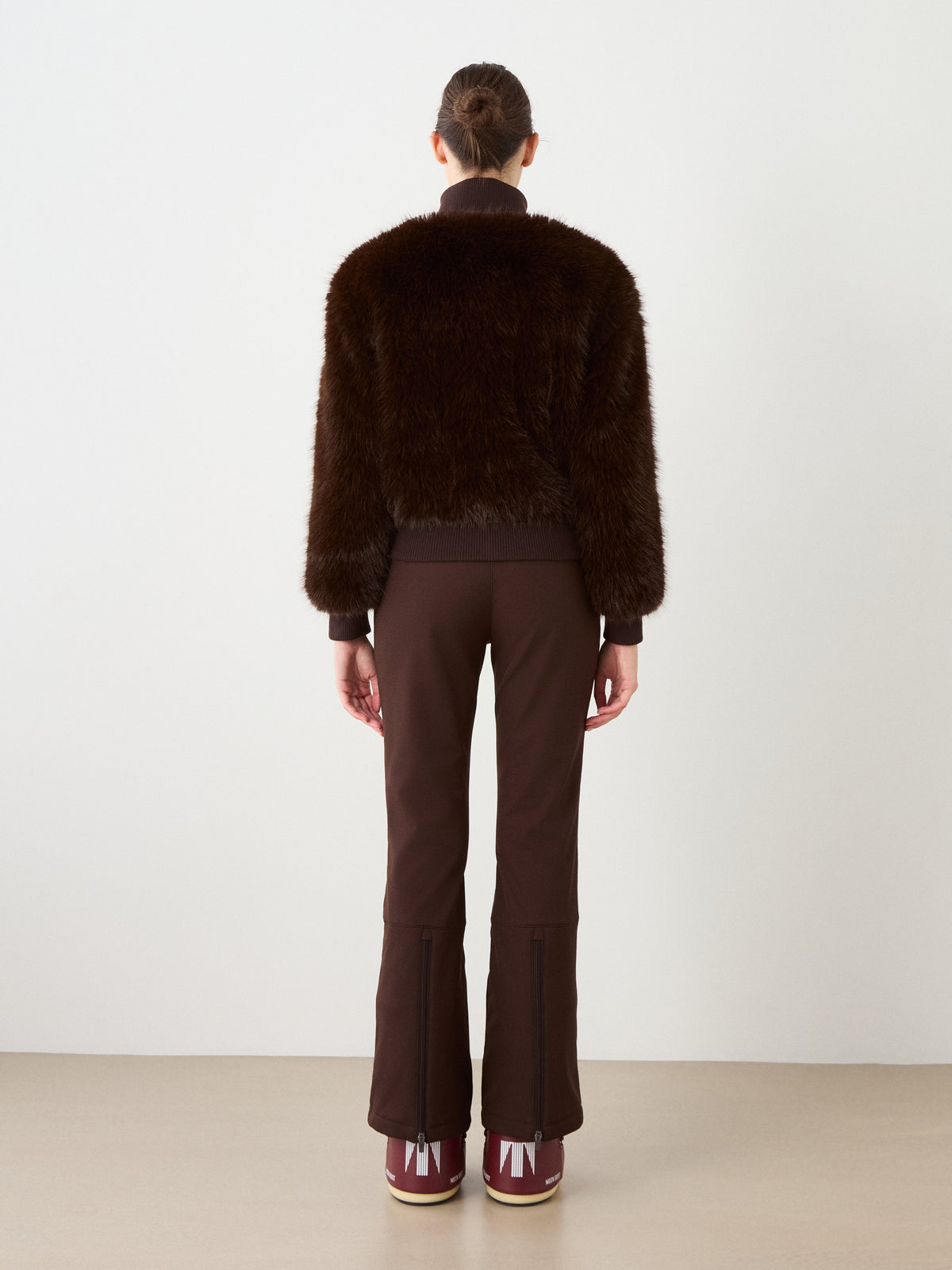Facing away from the camera, a person showcases effortless alpine style in the We Norwegians Dovre Jacket Women—a luxurious dark brown faux fur bomber—teamed with brown flared pants and maroon boots with crisp white details, set against a simple light background.