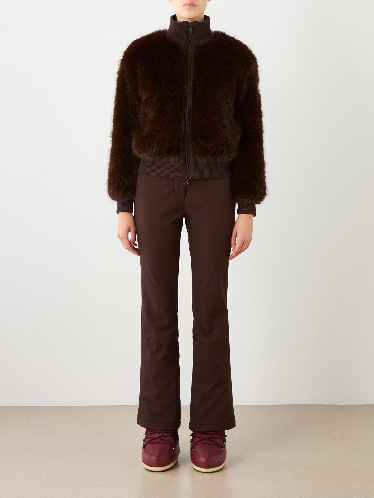 A woman stands against a plain background, wearing the We Norwegians Dovre Jacket Women—a dark brown faux fur bomber inspired by alpine style—paired with brown pants and burgundy boots. She faces forward with her arms relaxed at her sides.