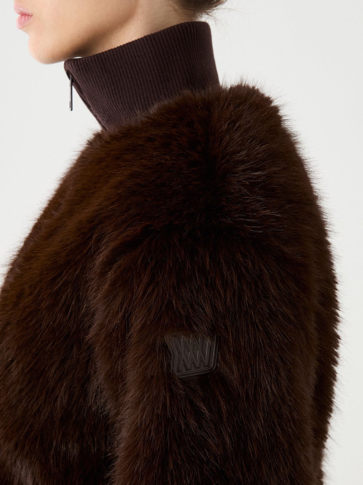 A person is wearing the We Norwegians Dovre Jacket Women—a dark brown faux fur bomber with a ribbed, high-neck collar. The image highlights the coat’s textured alpine style and a small patch on the sleeve, with only the lower half of the wearer’s face visible.